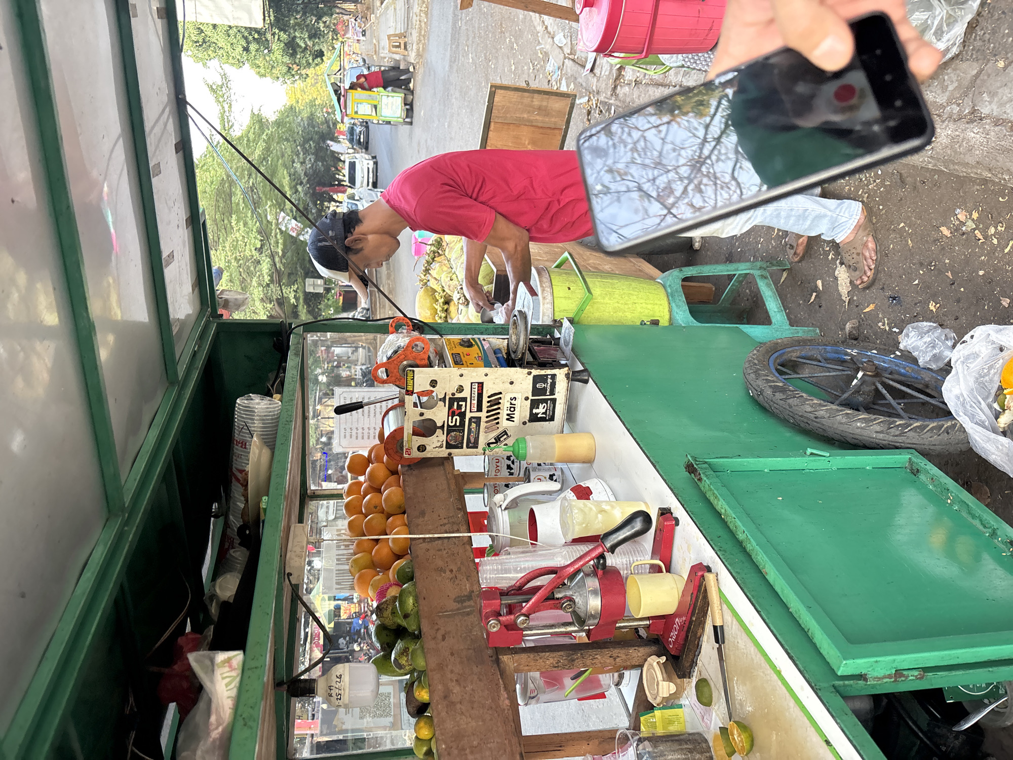 A street vendor in Cirebon, Indonesia, operates a fruit juice stand. The vendor, wearing a red shirt and a cap, is seen preparing a drink, with a smartphone in the foreground capturing the scene. The stand is well-stocked with fresh oranges, avocados, and limes. A manual juicer and various containers for liquids are visible. The background shows a typical Cirebon street scene with passing vehicles and other street vendors.