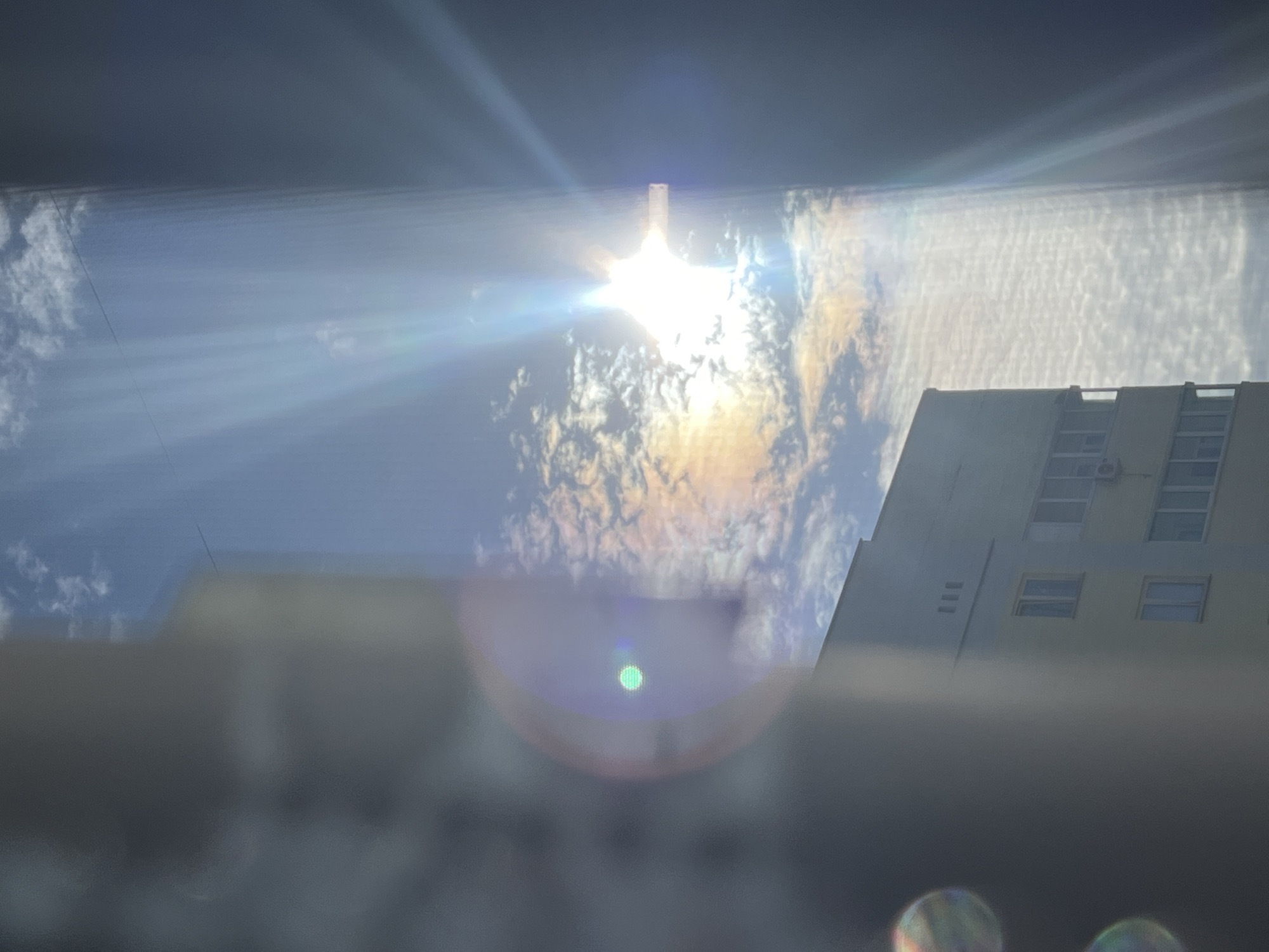 The image, taken in Chaikovsky, Russia, shows a bright sun with distinct rays of light extending downwards and to the right, partially obscured by clouds. A lens flare is visible, with a green circular reflection in the left portion of the image and two blurred, iridescent circular reflections in the lower left. In the lower half of the image, a light-colored building with multiple windows and visible air conditioning units is present. A dark vertical element, possibly a window frame or a building, occupies the right side of the frame, casting a shadow over the scene. The sky is blue with scattered white clouds.