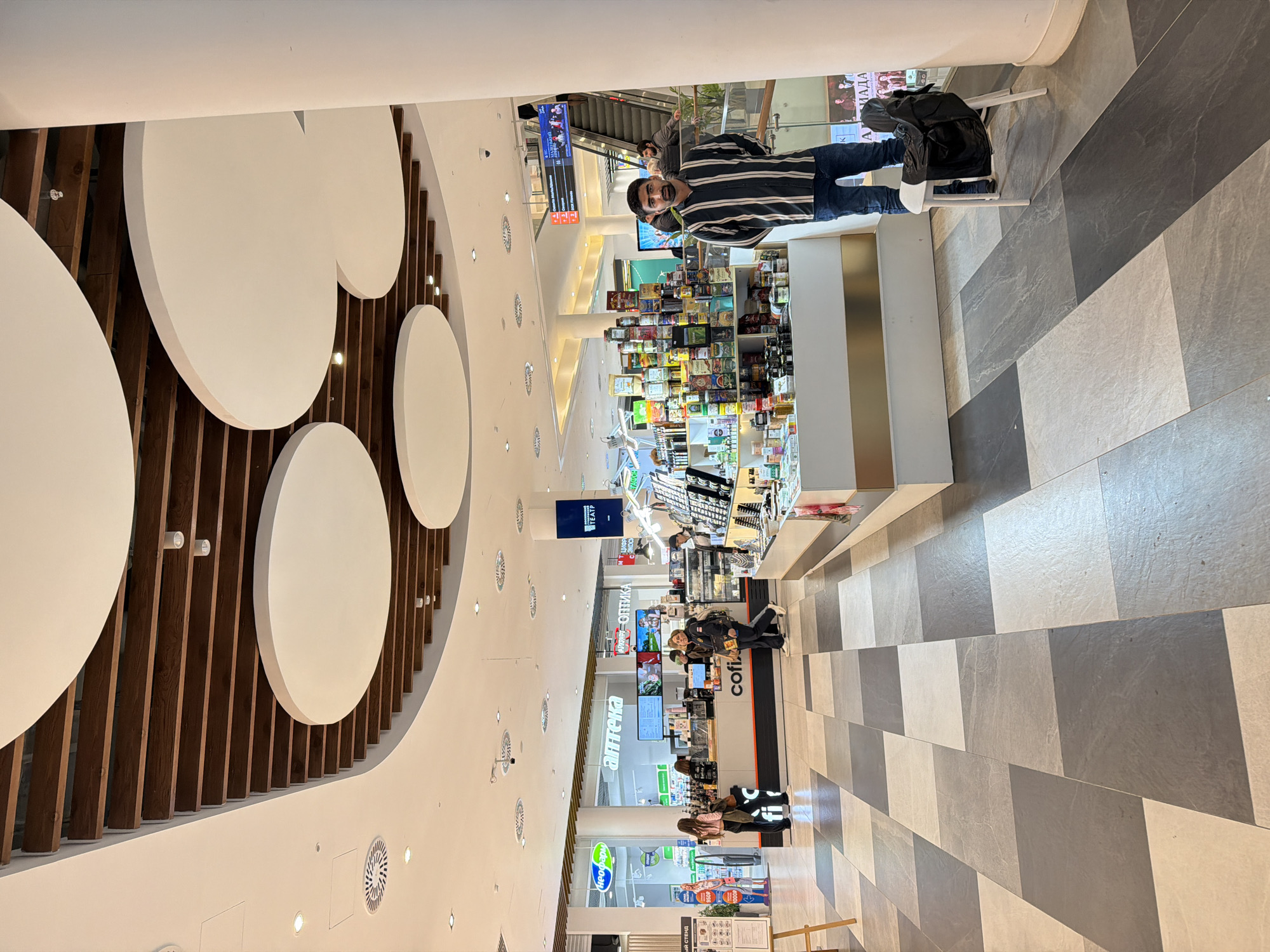 Here's a detailed description of the image:
**Location:** A shopping mall in Moscow, Russia. Evidence for this lies in the Cyrillic script on the storefronts ("аптека" is Russian for "pharmacy") and the visual style of the mall.
**Environment:** The mall has a modern, bright interior. The flooring is a patterned tile in alternating shades of grey and white. The ceiling is high, with a decorative design featuring large, white circular shapes and vertical wooden beams.
**People:**
* **One man is standing:** He is wearing a striped shirt, jeans, and sneakers. He is facing the camera with a slight smile. He has dark hair and a beard.
* **Other people are present in the background:** They are walking or standing near the shops, and are in a blurry state of detail.
**Objects:**
* **Shops:** Various retail stores line the mall's corridor, including a pharmacy and a café ("cofix").
* **Display cases:** There are tall display cases with goods stacked on shelves.
* **Seating:** There is a chair.
* **Bag:** The man is carrying a bag.
* **Digital Displays:** There are digital displays.
**Actions:**
* The man is posing for the photo.
* Other people are browsing in the mall.
**Interactions:**
* It is implied that the man is either with someone who is taking the picture or is taking a selfie.