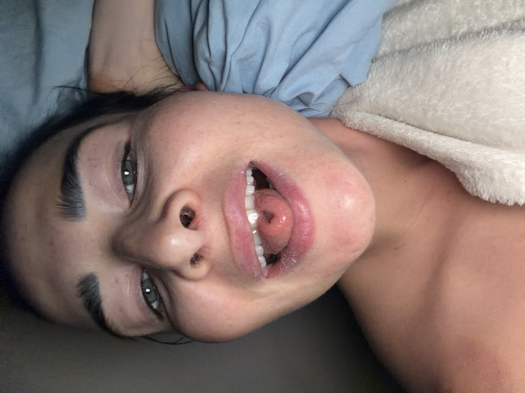 A close-up, angled shot depicts a woman's face. She is lying down, with her head tilted back. Her mouth is open, revealing her teeth and tongue. She has dark hair, fair skin, and noticeable eyebrows. Her eyes are open, and she is looking at the camera. A blue fabric or garment is draped behind her head, and a white, textured material is visible to the right of her shoulder. The background is out of focus and appears to be a solid, dark gray or brown. There is no location given.