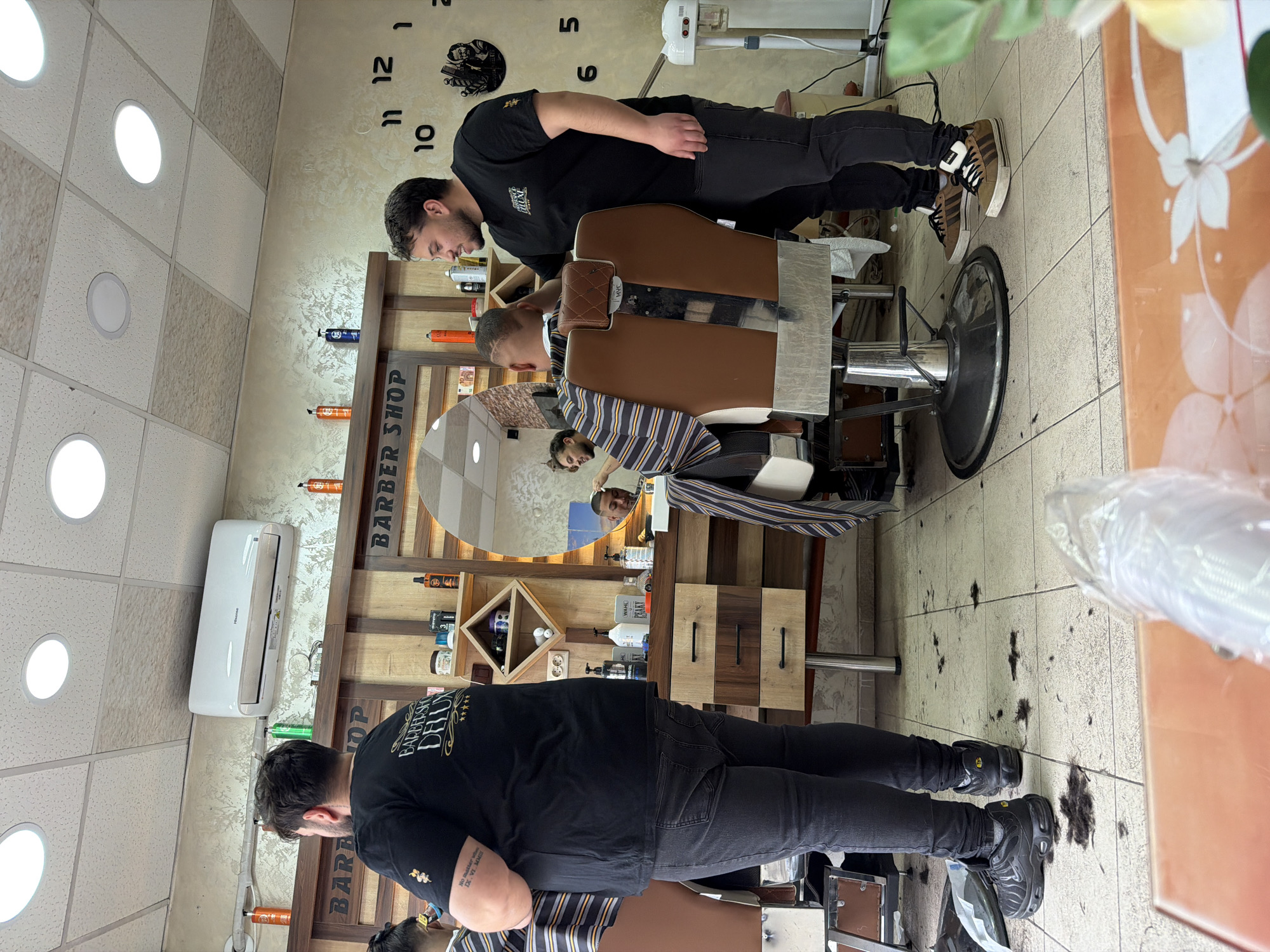 The image shows the interior of a barbershop. Two barbers, both wearing black shirts and dark jeans, are attending to two customers seated in barber chairs. One barber is actively cutting a customer's hair. A large mirror is visible behind one of the barbers, reflecting another customer receiving a haircut. The barbershop has a decorative clock on the wall and shelves displaying various hair care products. The floor is tiled, and there are cuttings of hair scattered on the floor. An air conditioning unit is mounted on the wall. The image appears to be taken indoors, likely in Panagyurishte, Bulgaria, although this location is not explicitly confirmed by visual elements.