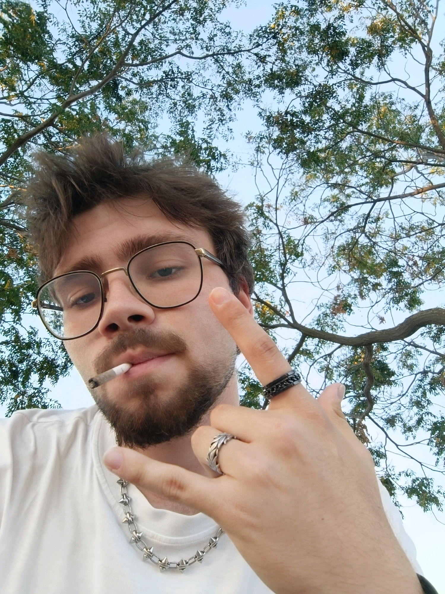 A young adult male with brown hair, glasses, and a beard is pictured outdoors. He is wearing a white t-shirt and a spiked chain necklace. He is holding a cigarette in his mouth and making a hand gesture. The background shows tree branches and a blue sky. The image is a close-up selfie taken from a low angle.