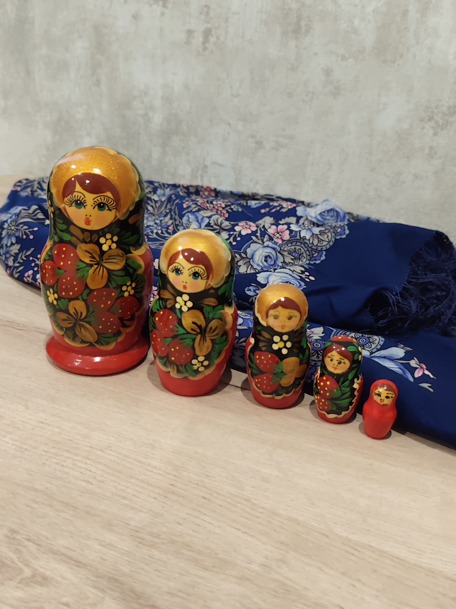 A wooden table has a stack of five Matryoshka dolls arranged from largest to smallest from left to right. The largest doll, on the far left, has a red base and a gold head with painted yellow, blue, and black features. Its body is decorated with strawberries and floral motifs in red, green, and yellow. The second largest doll shares a similar design. The third and fourth dolls are smaller and also feature the same decorative pattern. The smallest doll, on the far right, is entirely red and has minimal facial features painted in yellow. Behind the dolls, a folded dark blue scarf with a floral pattern in shades of pink, blue, and white is draped. The background consists of a textured gray wall. The image depicts an indoor setting with soft lighting.