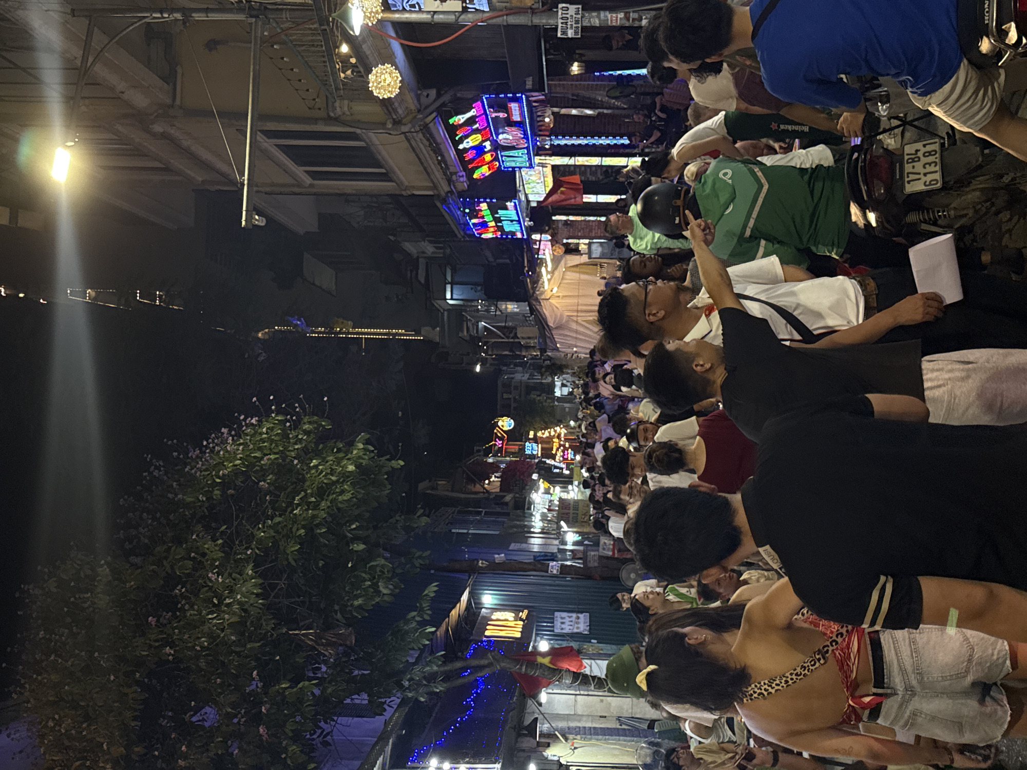The image depicts a crowded street scene at night in Hà Nội, Vietnam. Many people are present, some standing and others sitting. The street is illuminated by various sources of light, including streetlights and neon signs. A large tree is visible on the left side of the image. A motorbike with license plate 17-B4 613.81 is also visible.