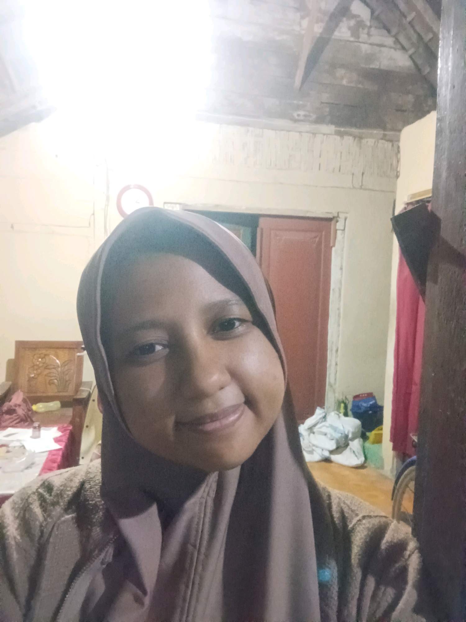 A medium shot captures a young woman in a brown hijab and a patterned cardigan, facing the camera from chest up, in an indoor setting. The woman has dark skin, brown eyes, and a subtle smile. Her hijab is a muted brown, and the cardigan has a textured, somewhat fuzzy appearance.
Behind the woman, a brown wooden door is visible, with a red frame. To the left of the door, a portion of a wooden chair with intricate carvings is seen, partially covered by a pink cloth. On a table to the left of the chair, there are several items including a bottle and what appears to be a container.
The background features a lightly colored wall with visible horizontal lines, possibly indicating wood paneling or peeling paint. Above this, a ceiling with exposed wooden beams and rafters is visible, some of which appear damaged or weathered. A circular object with a red border and a white center is mounted on the wall, to the upper left of the woman's head.
The lighting in the scene appears to be artificial and comes from above, creating bright areas on the ceiling and casting shadows. The overall environment suggests a dwelling, possibly in a rural or older building. The specific location of CANGA'AN, Indonesia is not explicitly indicated by any visual cues in the image itself.