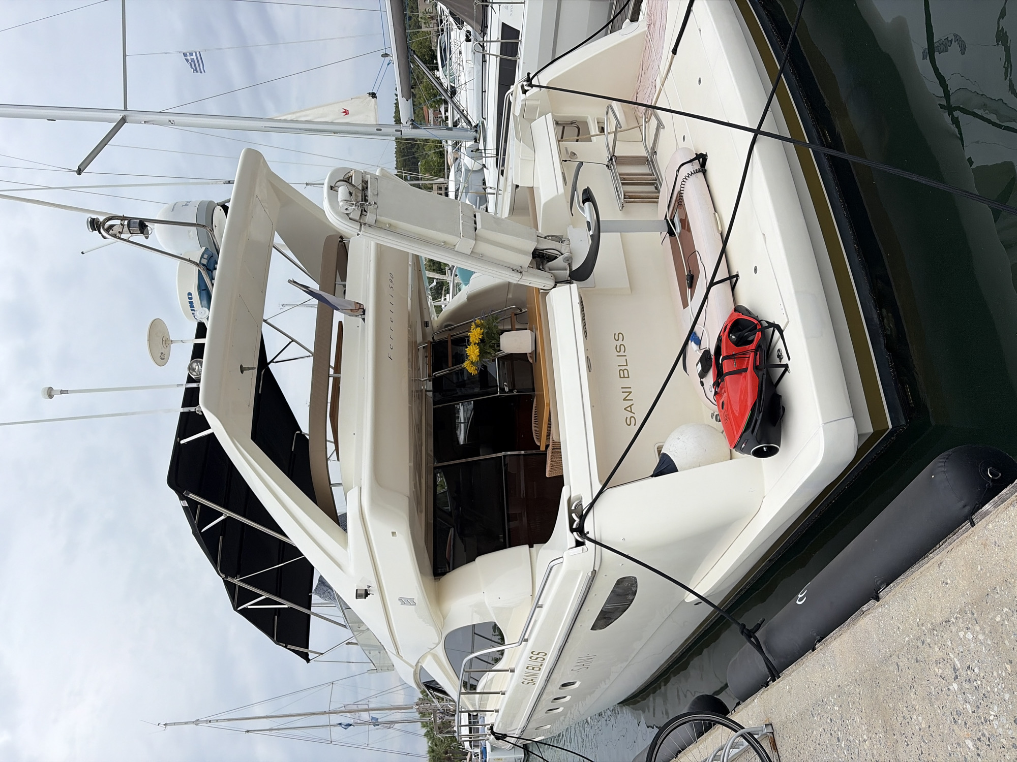 A white Ferretti 590 yacht named "SANI BLISS" is moored at a concrete dock. The yacht is viewed from the stern and port side. A black canopy is visible on the upper deck, supported by stainless steel poles. A davit arm extends upwards from the stern. On the deck, a red submersible drone and a white board are present. The water is dark and reflective, with visible mooring lines. In the background, other boats and masts are seen, along with a glimpse of green foliage and buildings on a distant shore under a cloudy sky. The Greek flag is flying from a flagpole on a nearby vessel. The location appears to be a marina in Greece, likely in Kassandra Municipal Unit, given the Greek flag and coastal setting. No people are visible in the image.
