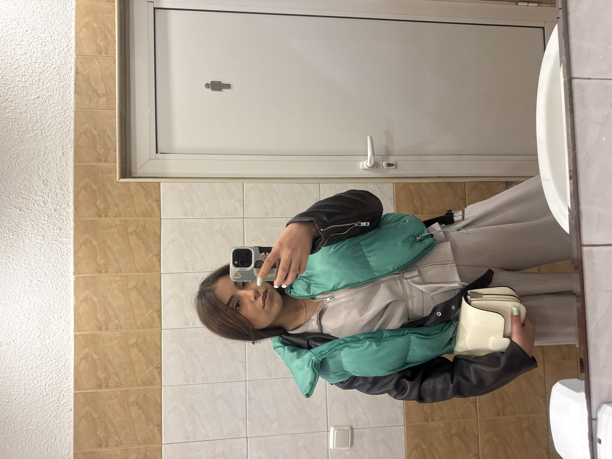 A young woman is taking a selfie in a restroom. The restroom features beige and white tiled walls, a white door with a gender symbol, and a white sink partially visible on the right. The woman wears a black jacket with a green puffy vest, a light-colored outfit underneath, and holds a small white purse. She is using an iPhone to capture the selfie. The location is potentially in Sofia, Bulgaria, but there is no verifiable information in the image to confirm this, making it an assumption based on prior knowledge.