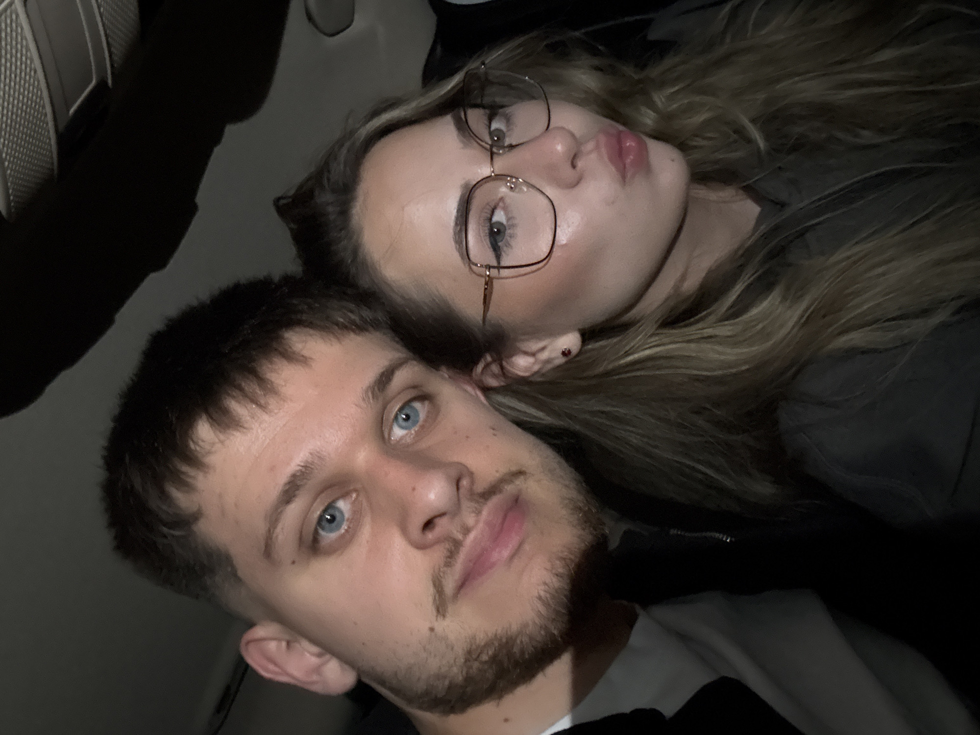 A dimly lit selfie shows a young man and woman. The man is in the foreground, his face angled toward the camera. He has short, dark hair, blue eyes, and stubble on his face. The woman is positioned behind him, lying down with her head slightly raised. She has long, blonde hair, wears glasses, and her lips are pursed. The background is dark and indistinct, suggesting the photo was taken indoors, possibly inside a car. There is no indication of Blagoevgrad, Bulgaria.