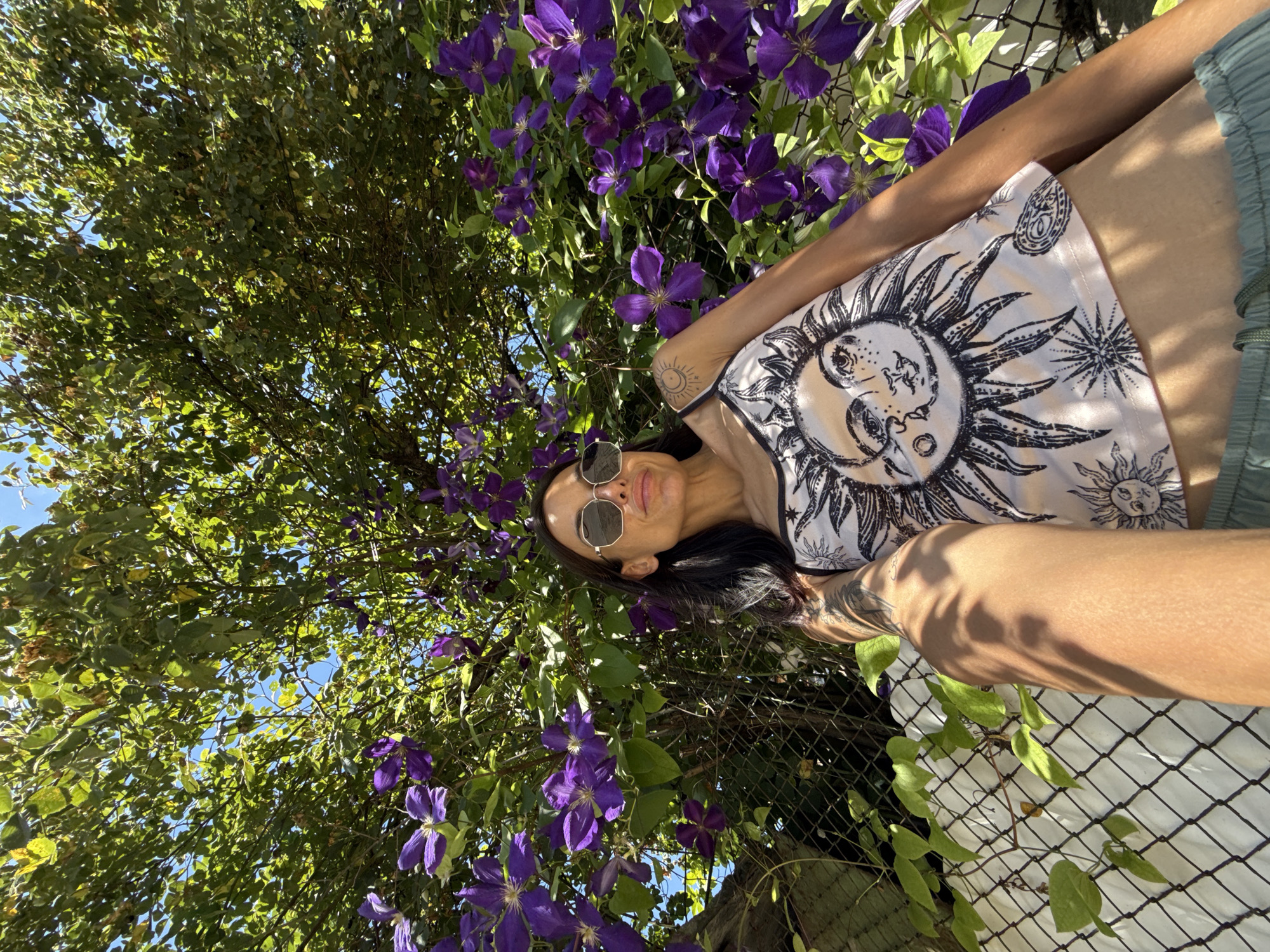 In a sun-drenched outdoor setting, a woman with dark hair and sunglasses poses beneath a canopy of lush green leaves and vibrant purple clematis flowers. She is wearing a light-colored tank top adorned with a black sun graphic and light green shorts. Tattoos are visible on her left arm and shoulder. The background features a chain-link fence partially covered with a white tarp, and behind it, more foliage and a clear blue sky. The scene appears to be a private garden or yard.