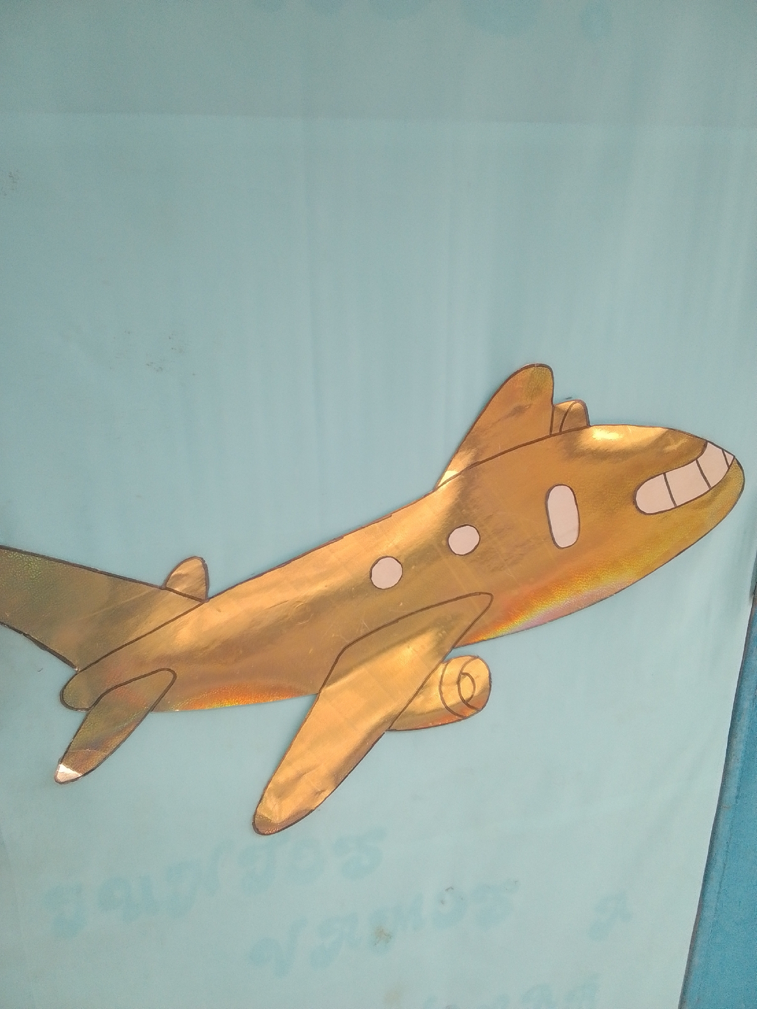 A metallic gold cutout of an airplane is displayed against a light blue backdrop. The airplane is depicted in profile, facing to the right, with a streamlined body, swept-back wings, and two engines mounted beneath the wings. It has two oval windows and a cockpit area with two simple windows resembling teeth. The airplane's surface has a shiny, iridescent quality, reflecting rainbow-like colors in places. The backdrop appears to be a fabric banner or wall, with subtle vertical creases suggesting it might be a large piece of material. Faint, blurred text is visible at the bottom of the image, hinting at a possible inscription or title, though it is illegible. There are no people visible in the image, and the focus is entirely on the airplane cutout and the backdrop. The lighting suggests an indoor setting or an overcast outdoor environment, as there are no strong shadows. The overall impression is that of a decorative element, possibly for a classroom, event, or children's area in La Tagua, Venezuela.