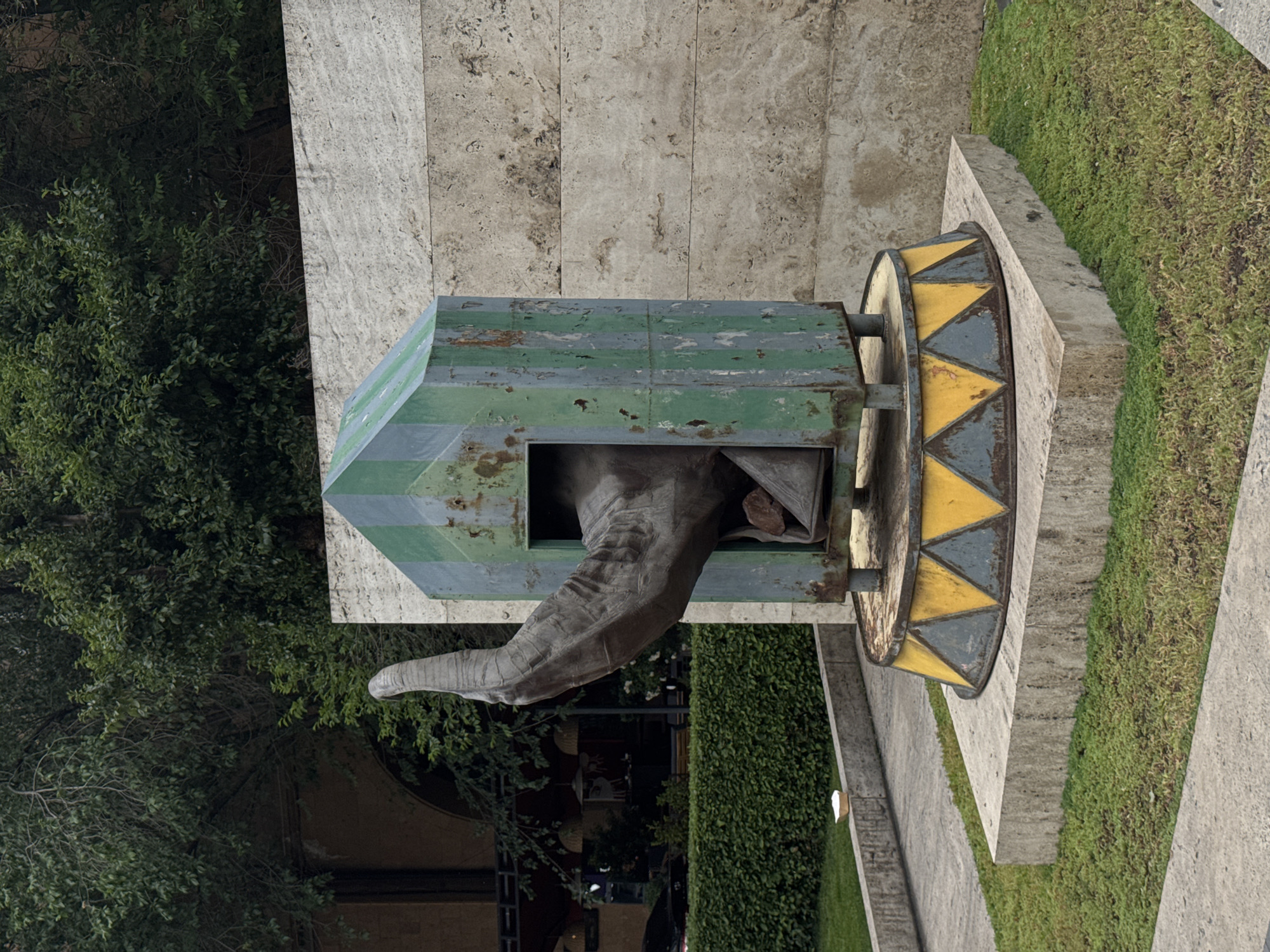 The image depicts a modern art installation in Yerevan, Armenia. It consists of a green and gray striped box-like structure with an opening from which a large, weathered-looking sculpted hand extends. Attached to the back of the box is a circular element decorated with alternating yellow and gray triangles. The installation is situated on a light-colored concrete base surrounded by grass and foliage. In the background, there are trees, a building with a stone facade, and a partial view of what appears to be a street or pathway with some indistinct figures.
