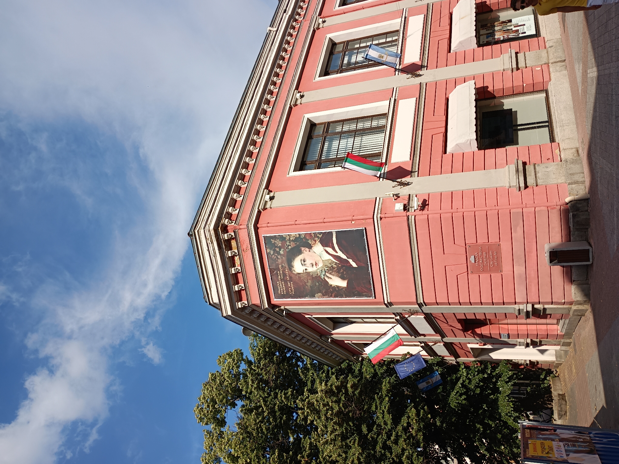 This is a photograph of a pink building with white trim, likely an art gallery. The building is adorned with several flags, including the Bulgarian flag and what appears to be the European Union flag. A large portrait of a young woman in historical attire is prominently displayed on the facade. The image is taken from a low angle, looking upwards towards the building and the sky, which is blue with scattered white clouds. A large tree with green foliage is visible in the foreground, partially obscuring the lower part of the building. The scene suggests a sunny day. Text visible on the building includes "ГРАДСКА ХУДОЖЕСТВЕНА ГАЛЕРИЯ - ПЛОВДИВ CITY ART GALLERY PLOVDIV" and "ГРАДСКА ХУДОЖЕСТВЕНА ГАЛЕРИЯ City Gallery of Fine Art", confirming the location as the City Art Gallery in Plovdiv, Bulgaria. A partial view of a person wearing a yellow shirt is visible at the top right of the frame. There is also some text on posters at the bottom of the image, but it is mostly illegible.