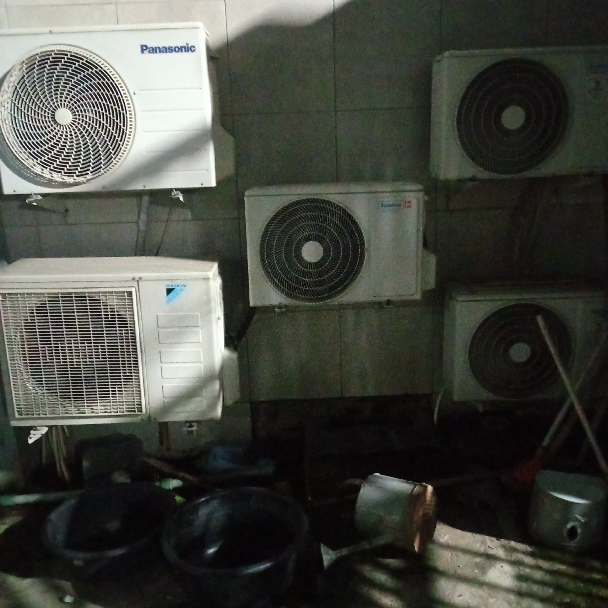 The image shows an outdoor scene with multiple air conditioning units mounted on a tiled wall. The units appear to be in a residential or commercial area, possibly a back alley or utility area, given the presence of several large black basins and cleaning tools in the foreground, including a broom and dustpan. The lighting suggests it is either nighttime or a dimly lit indoor space. One air conditioning unit on the top left is clearly labeled "Panasonic". Another unit to its right is partially visible and appears to be a different brand. Below the Panasonic unit, on the left, is a Daikin unit. To the right of the Daikin unit are two more air conditioning units, one above the other. The overall impression is one of a functional but somewhat cluttered outdoor space, with the air conditioning units being the primary focus. There is no indication of the specific location within Ibadan, Nigeria, beyond what can be inferred from the general context of the scene.