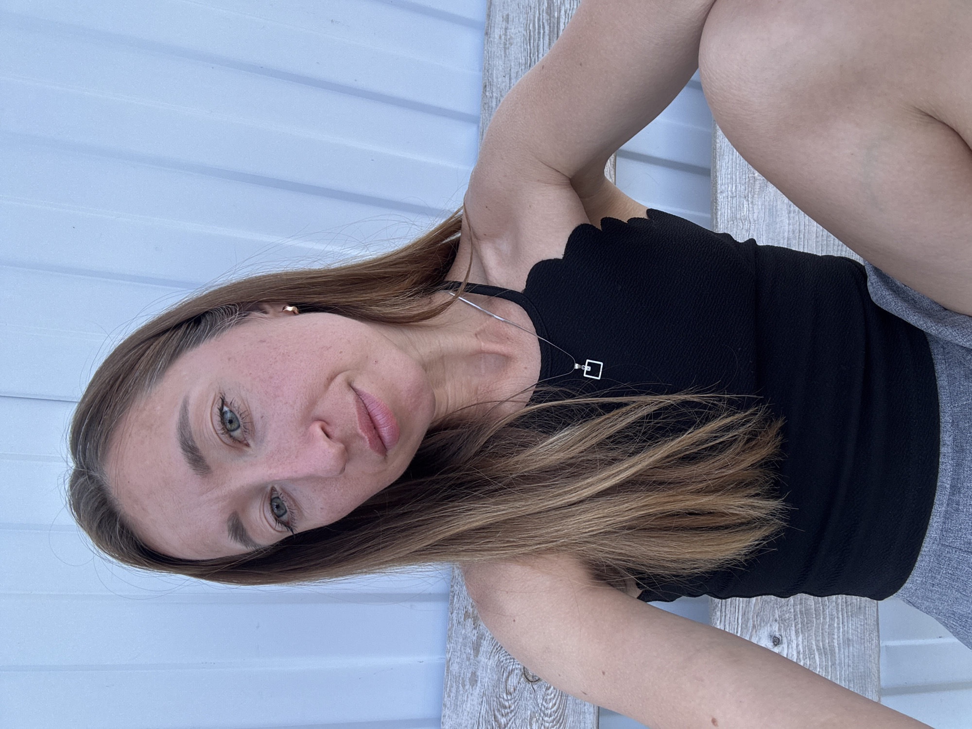 A woman with long, light brown hair, green eyes, and fair skin is sitting outdoors. She's wearing a black top with a scalloped neckline and a silver necklace with a square pendant. She is sitting on a light-colored wooden structure, possibly a bench or step. She is also wearing gray pants. Behind her is a white wall with horizontal paneling. The location in the city of Chaikovsky, Russia is not determinable from the given image.