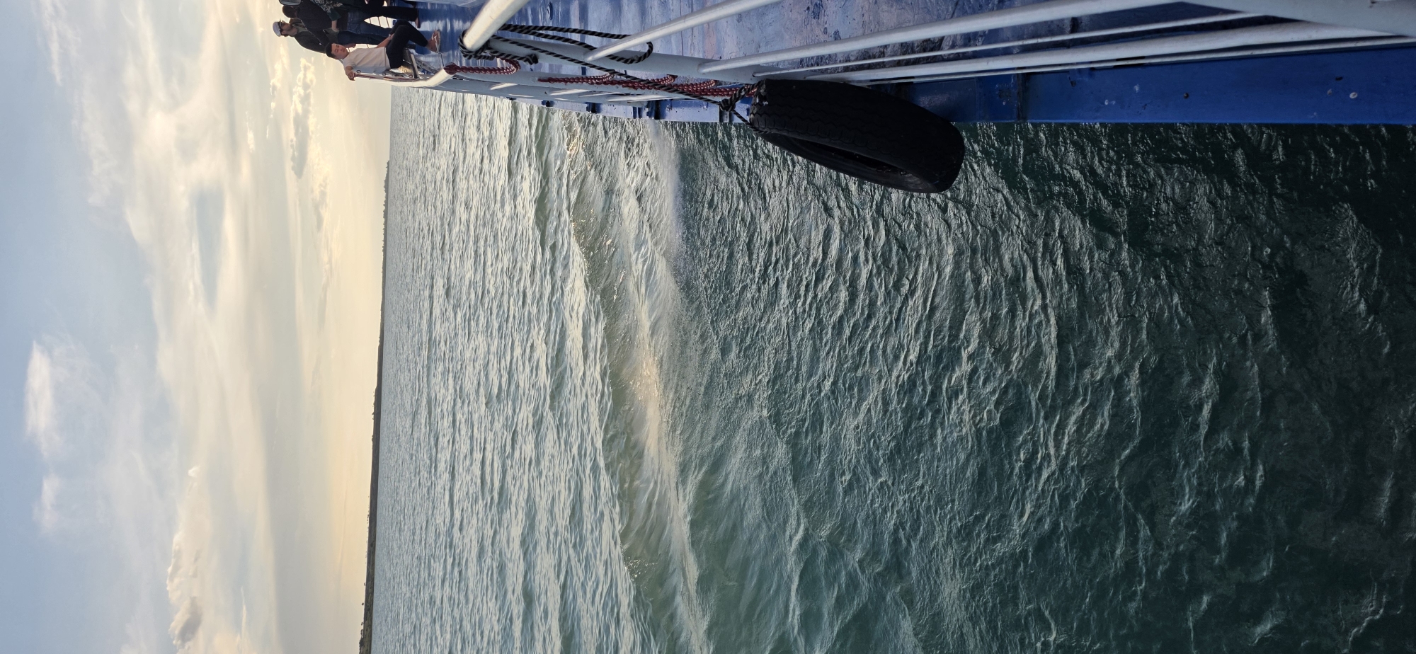 The image depicts a view from the side of a boat on a body of water, likely a lake or sea, given the distant landmass visible on the horizon. The water is choppy, with small waves and ripples reflecting the overcast sky. The sky itself is filled with scattered clouds, with a soft, diffused light suggesting it might be early morning or late afternoon.
On the right side of the frame, the blue railing of the boat is visible, with a black tire fender hanging from it. A rope with red and black accents is also attached to the railing. Several individuals, appearing to be passengers, are standing or sitting along the railing, looking out at the water or the horizon. One person in the foreground is leaning on the railing, dressed in casual attire. Further back, other figures are silhouetted against the sky. The boat appears to be moving, as indicated by the wake in the water.
The environment is serene and expansive, characterized by the vastness of the water and the sky. The distant land appears as a low-lying strip, possibly a shoreline with sparse vegetation. The overall impression is one of a peaceful journey on the water under a cloudy sky. The specific location in Pristan'-Przheval'sk, Kyrgyzstan, implies this body of water is likely Issyk-Kul Lake, a prominent natural attraction in the region.