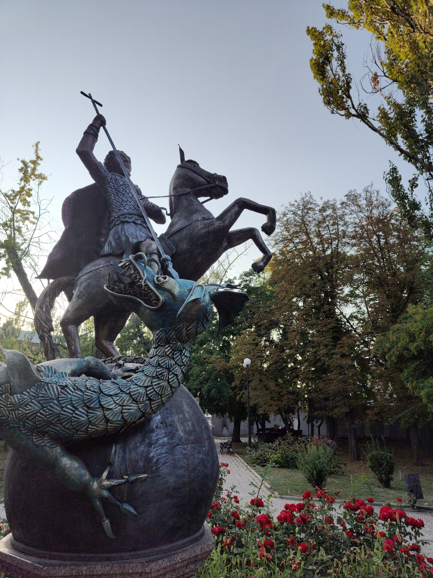 A bronze statue of Saint George slaying a dragon stands in a park in Donetsk, Ukraine. The statue depicts Saint George on horseback, spearing the dragon, which is coiled around a sphere. The dragon has a scaly body and sharp claws. The statue is located in front of a path lined with red roses. In the background, trees and a lamppost are visible. The sky is clear and blue.