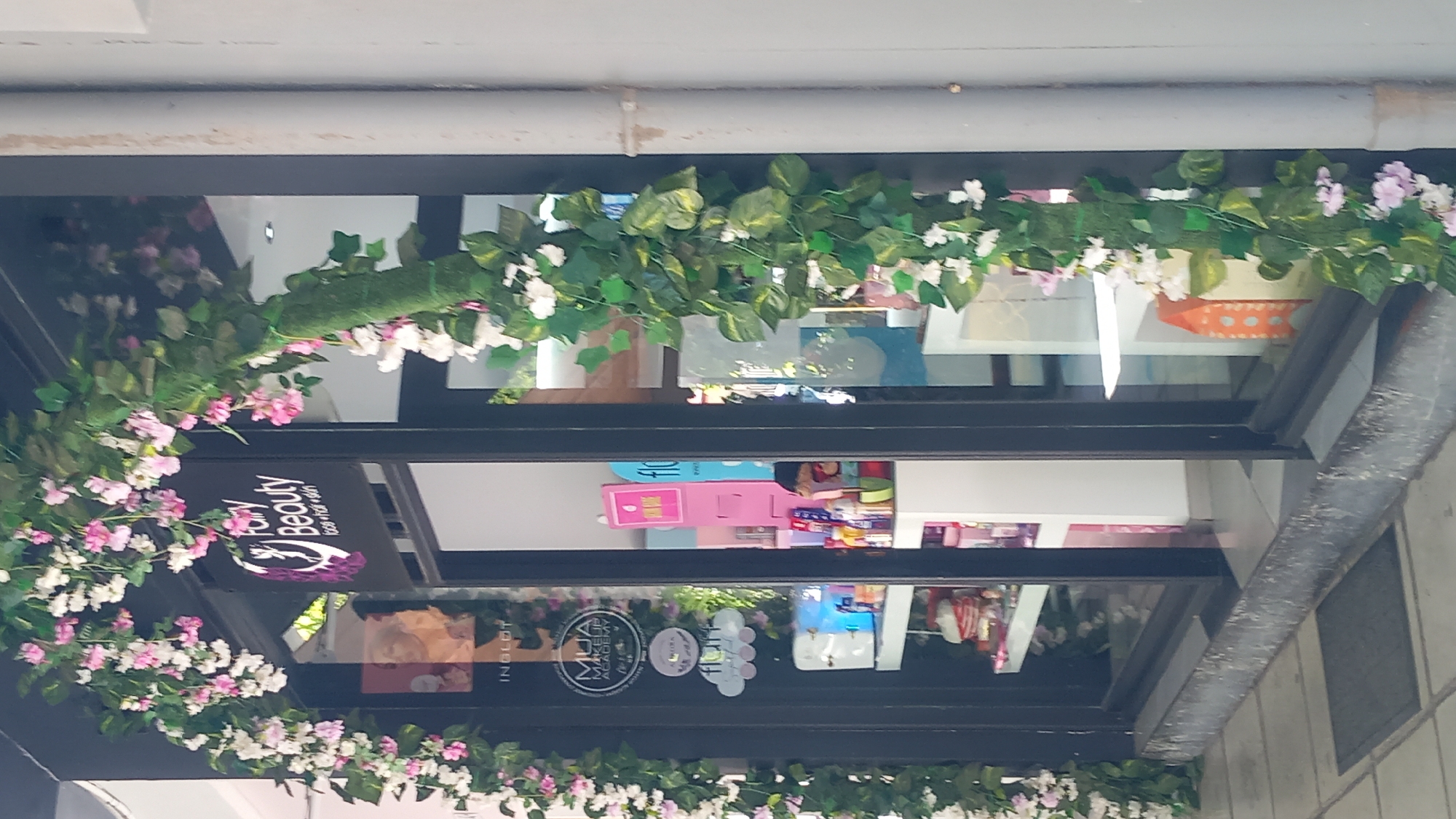 The image shows the storefront of a beauty salon named "Fairy Beauty" in Kos, Greece. The entrance is decorated with an archway of artificial flowers and greenery, predominantly pink and white blossoms with green leaves. The salon's name and slogan "face • hair • skin" are visible on a sign inside the window. Through the glass, various beauty products are displayed on shelves, and some display stands are visible. The exterior of the building appears to be modern, with dark-framed glass windows and doors. The ground in front of the shop is paved with gray tiles, and a dark doormat is placed outside the entrance. The overall impression is of a well-maintained and visually appealing commercial establishment.