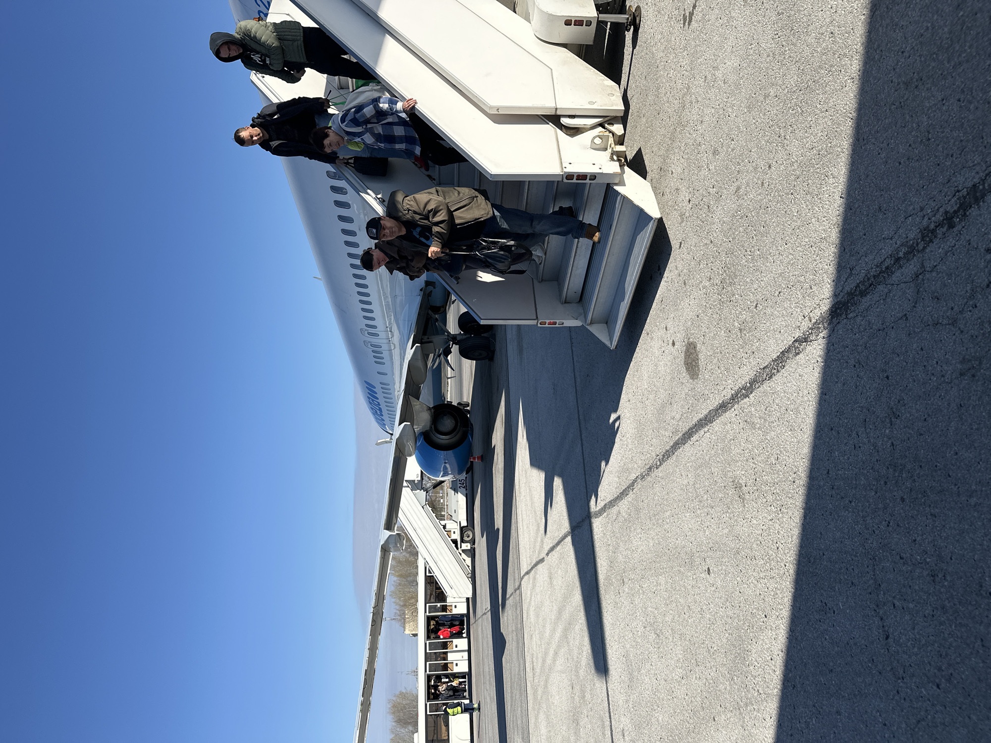 People are deplaning from an airplane at Bolshoye Savino Airport in Bolshoye Savino, Russia. Passengers are descending the stairs from the airplane onto the tarmac. The aircraft is connected to a mobile passenger boarding stair truck, located at the bottom of the stairs. A ground crew person is standing near the truck. The sky is clear and blue.