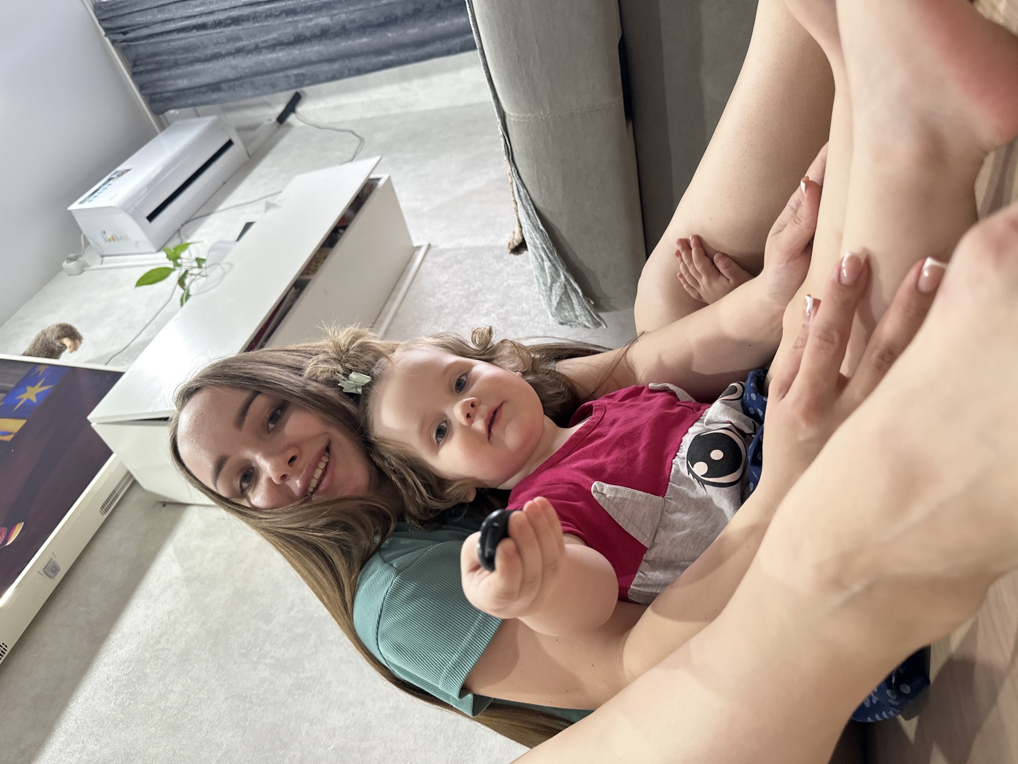 A woman is holding a young child. The woman has long brown hair, fair skin, and is wearing a green top. The child has light brown hair, fair skin, and is wearing a red top with gray sleeves. The child is holding a small black object in their right hand. The woman is smiling at the camera. The background includes a white cabinet, an air conditioning unit, and part of a gray couch. The location is a domestic setting, potentially a home in Chaikovsky, Russia. The interaction between the woman and child suggests a familial bond.