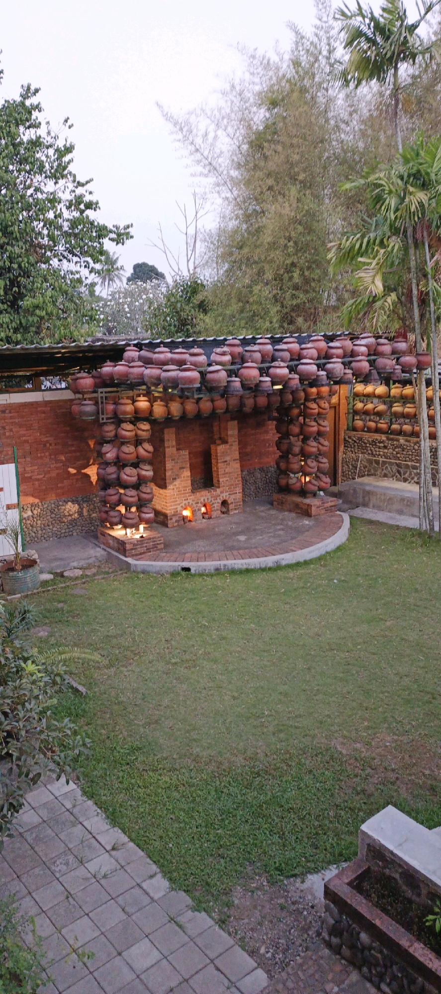 In the city of Sidokarto, Indonesia, this image captures an outdoor scene featuring a brick structure that functions as a pottery kiln and display area. The kiln itself is built from red bricks and has two arched openings in the front, with visible flames inside suggesting active use. Above and around the kiln, numerous terracotta pots are stacked and arranged. These pots form the roof and decorative elements of an open-sided structure connected to the kiln, creating an arched walkway. The pots are of various sizes and shapes, with some appearing to be traditional clay urns.
The area in front of the kiln is paved with bricks in a semi-circular pattern, creating a small patio. To the left of the kiln, a stone wall and a brick wall are visible, with more pots displayed on shelves attached to the brick wall. Green grass covers the ground in the foreground and middle ground, with some greenery and bushes to the left. In the background, tall bamboo trees and palm trees are visible against a pale sky, indicating a tropical or subtropical environment. A paved walkway made of square stone tiles runs along the bottom left of the image. There are no people visible in the frame. The overall impression is that of a functional artistic space dedicated to pottery, set within a garden or natural landscape.