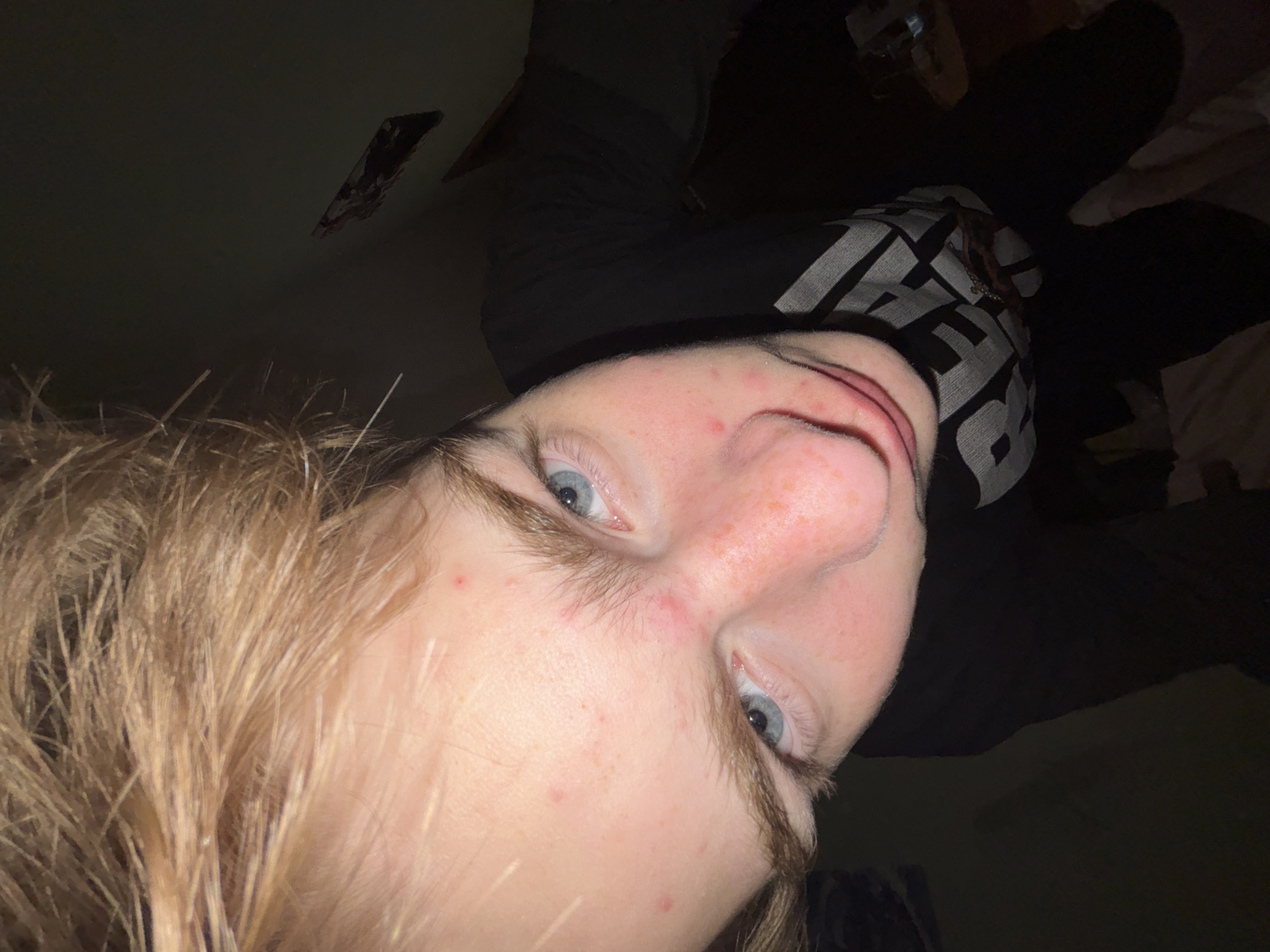 A close-up selfie of a person with light skin, blonde hair, and blue eyes. The person is wearing a dark shirt with white text on it. The background is dark and indistinct, with what appears to be a piece of paper or cardboard attached to the ceiling. The image is rotated, with the person's face angled towards the lower left corner. The location is Unknown, Unknown.