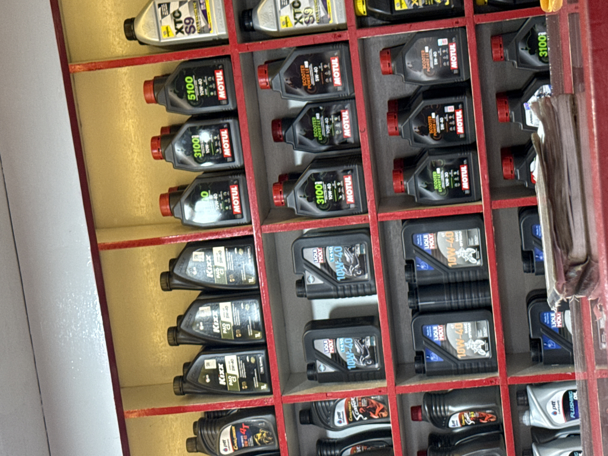 The image shows a display of various motor oils in a shop, likely in Lhokseumawe, Indonesia. The oils are arranged in a grid-like shelf system, with brands such as Motul, Kixx, Liqui Moly, and PTT visible. The Motul oils include variants like 3100, 5100, Scooter Power LE, and Scooter Expert LE. The Liqui Moly oils are labeled "T410 10W-40" and "Basic Offroad 10W-40." The PTT oils include "Challenger 4T" and "Flushing Oil." The setting appears to be a retail environment specializing in automotive products. The shelves are constructed with red framing.