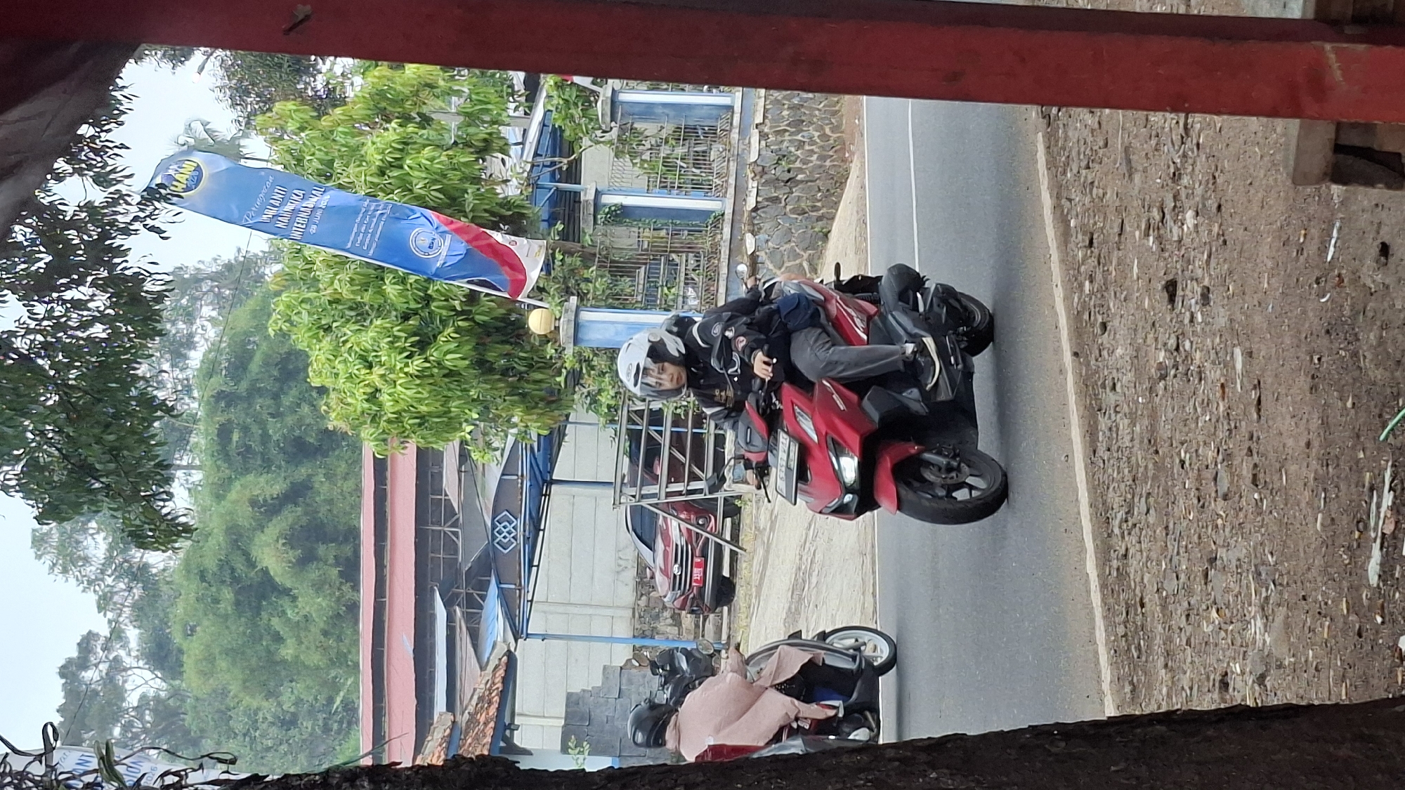 In Cilaku, Indonesia, a woman wearing a white helmet and dark jacket rides a red scooter. She is on a paved road, with trees and buildings in the background. Another person on a scooter, wearing a pink garment and a dark helmet, is visible to the left, further down the road. A red car is parked to the left of the road. A blue and white banner is hanging from a pole on the right side of the image. The sky is overcast.