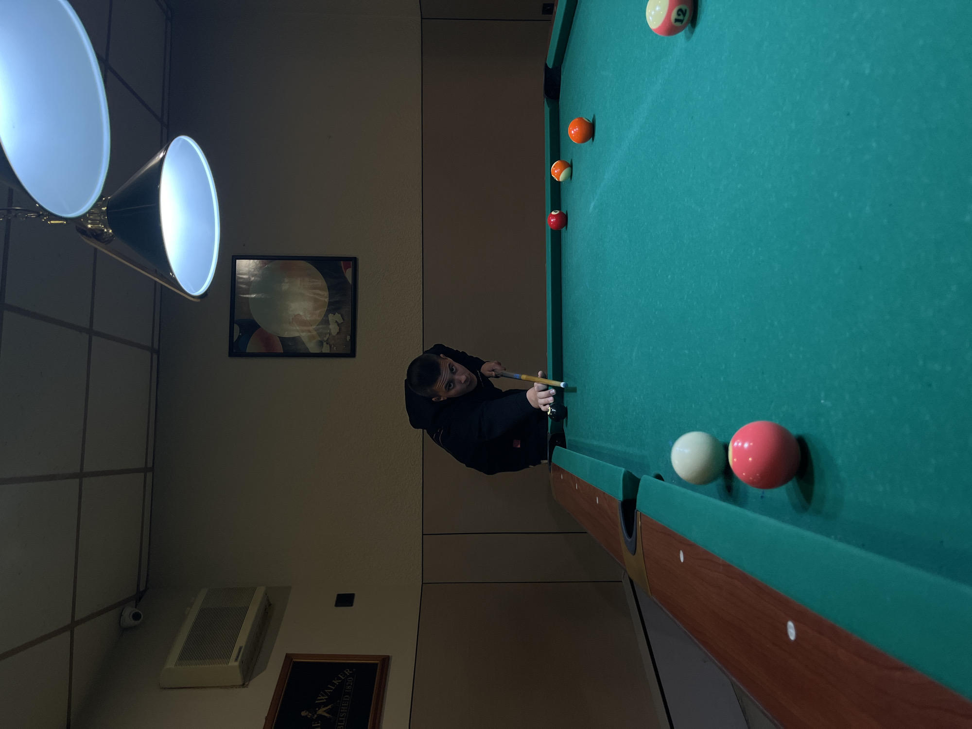 An indoor recreational facility, possibly a pool hall, in Berkovitsa, Bulgaria, is depicted. A male, appearing to be a young adult, is positioned over a green-felted pool table, preparing to execute a shot. He wears a dark-colored hooded sweatshirt and holds a wooden cue stick with his right hand while his left hand forms a bridge on the table surface. His gaze is directed forward, aligning the cue stick with the white cue ball positioned near the table's edge.
On the table, several colored billiard balls are visible: a solid red ball, a striped orange ball, a solid orange ball, a light pink/red solid ball, and a partially visible striped yellow ball with the number "12."
The room's walls feature a two-toned design, with a darker brown wainscoting effect on the lower half and a lighter beige paint above. A framed advertisement, partially legible as "Johnnie Walker, ESTABLISHED 1820," hangs on the left wall. Further right, an abstract framed artwork featuring circular motifs is mounted. The ceiling is a suspended grid with square panels. Two pendant lamps with dark conical shades and bright white interiors illuminate the pool table from above. A white wall-mounted air conditioning unit is visible on the upper left wall, beneath a dome-shaped security camera mounted on the ceiling, directed into the room.