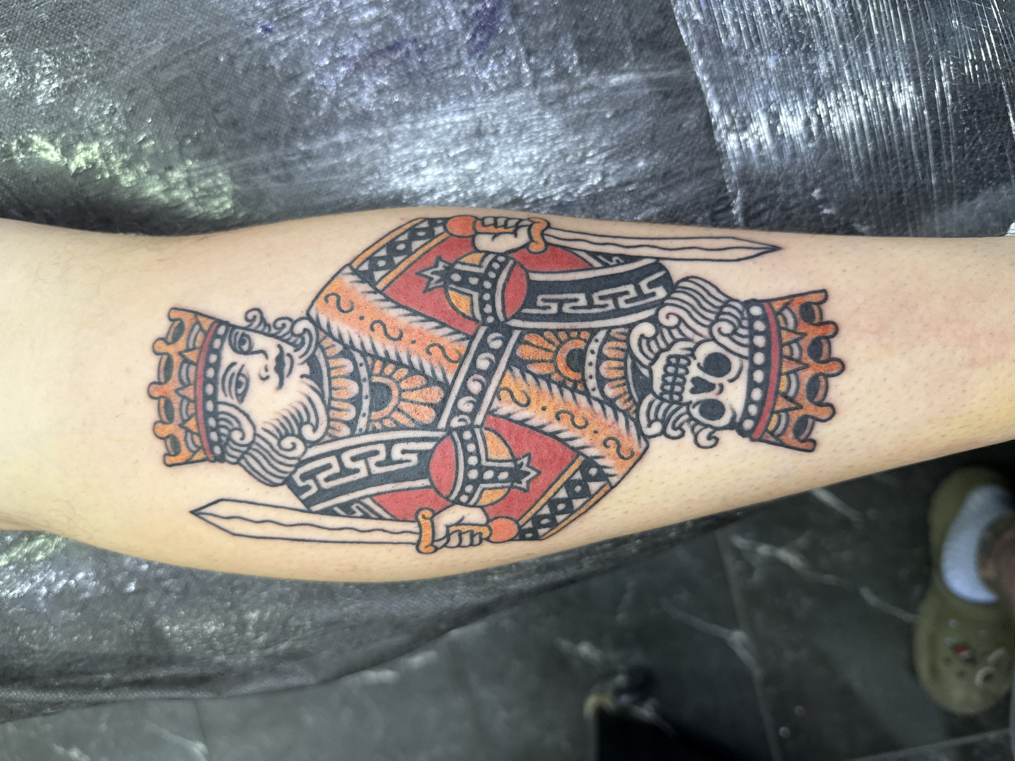 This close-up image displays a fresh, vibrantly colored tattoo on a person's leg or arm, likely the calf, with subtle redness indicating recent completion. The tattoo features a symmetrical design reminiscent of a playing card king. One half depicts a crowned king with a human face, beard, and flowing hair, adorned in ornate robes and holding a sword. The other half mirrors this design but instead shows a crowned skull, symbolizing a skeletal king, also holding a sword.
The tattoo is executed in a traditional American/European style, characterized by bold black outlines and solid color fills. The dominant colors are rich red, warm orange-gold, and black, with accents of off-white. The background consists of a dark, slightly reflective surface, possibly a floor or table, with some lighter textures visible. In the bottom right corner, a portion of a green Croc-style shoe is visible, suggesting a casual indoor setting, possibly a tattoo studio in Moscow, Russia. There is no discernible text within the image.