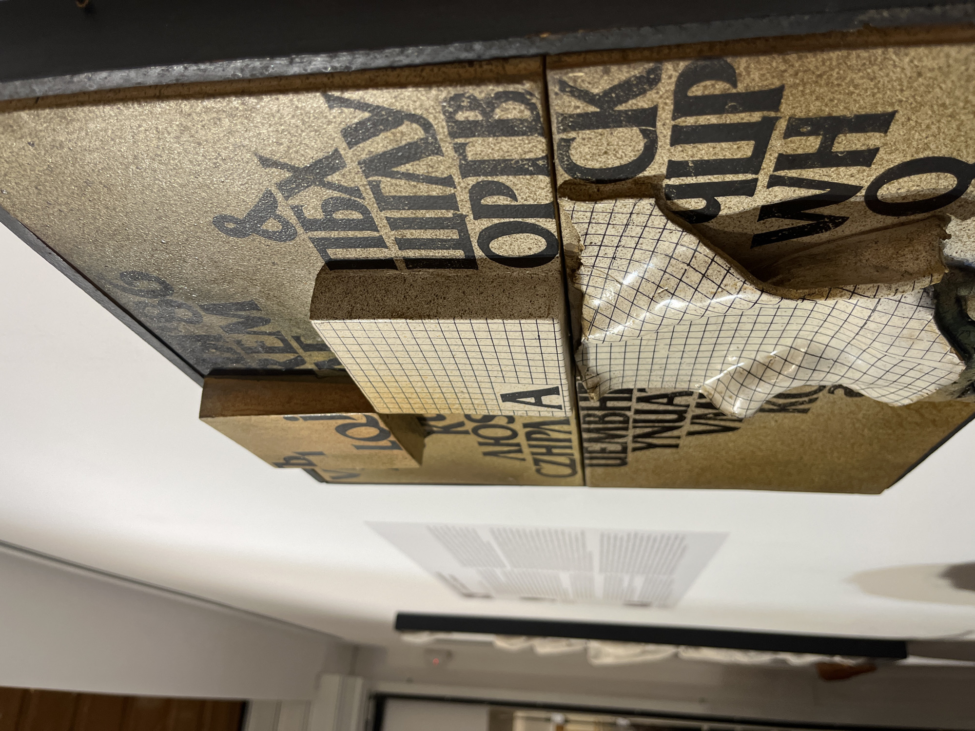 The image presents a close-up, upward-angled view of a mixed-media artwork or installation, likely displayed in an exhibition space in Plovdiv, Bulgaria. The piece is a three-dimensional relief, constructed from various rectangular blocks of material, possibly wood or particle board, mounted against a lighter background.
The surface of the artwork is predominantly a textured, sandy-beige color with bold, black, sans-serif letters printed upon it. Due to the artwork's orientation and the camera's perspective, much of the text appears inverted. Clearly visible characters and partial words include "&", "IBX", "OPTR", "CK", and "W H O". Below these, other visible letters and fragments include "LO", "A", "CZPA", "UEMBH", "YNUA", and "VA".
A distinctive feature is the application of a material resembling graph paper or a fine grid pattern onto some of the protruding blocks. This grid-patterned material is visibly peeling away in several areas, revealing the underlying structure and adding a sense of deconstruction or layered history to the piece. The installation is framed by dark, possibly black, borders at its top and bottom edges. The lighting emphasizes the artwork's three-dimensionality, casting shadows that highlight its depth and varied surfaces. The background, blurred, suggests an indoor setting, possibly an art gallery or museum, with faint indications of other displayed items or architectural details. No specific activity or people are depicted beyond the implied viewing of the art.