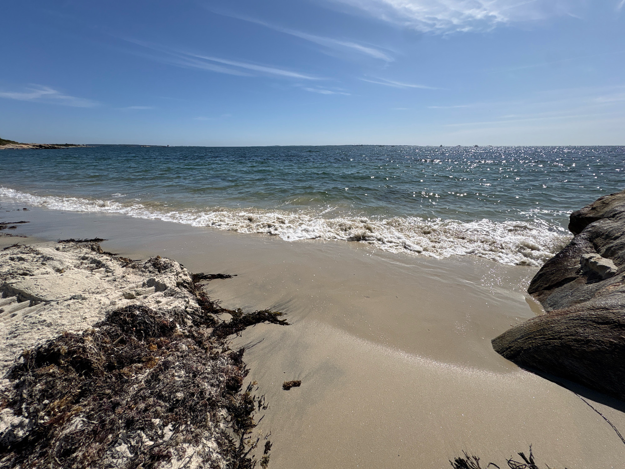 The image depicts a serene beach scene in Waterford, United States of America, under a clear blue sky dotted with wispy white clouds. The foreground is dominated by a sandy beach with a scattering of dark seaweed on the left side and a large, dark boulder on the right. The gentle waves of the ocean are seen lapping at the shore, their surface shimmering with sunlight. In the distance, the ocean stretches out to the horizon, where a few boats and what appear to be distant landmasses or structures are visible. The overall impression is one of a tranquil coastal landscape on a bright, sunny day. There are no people visible in the image.