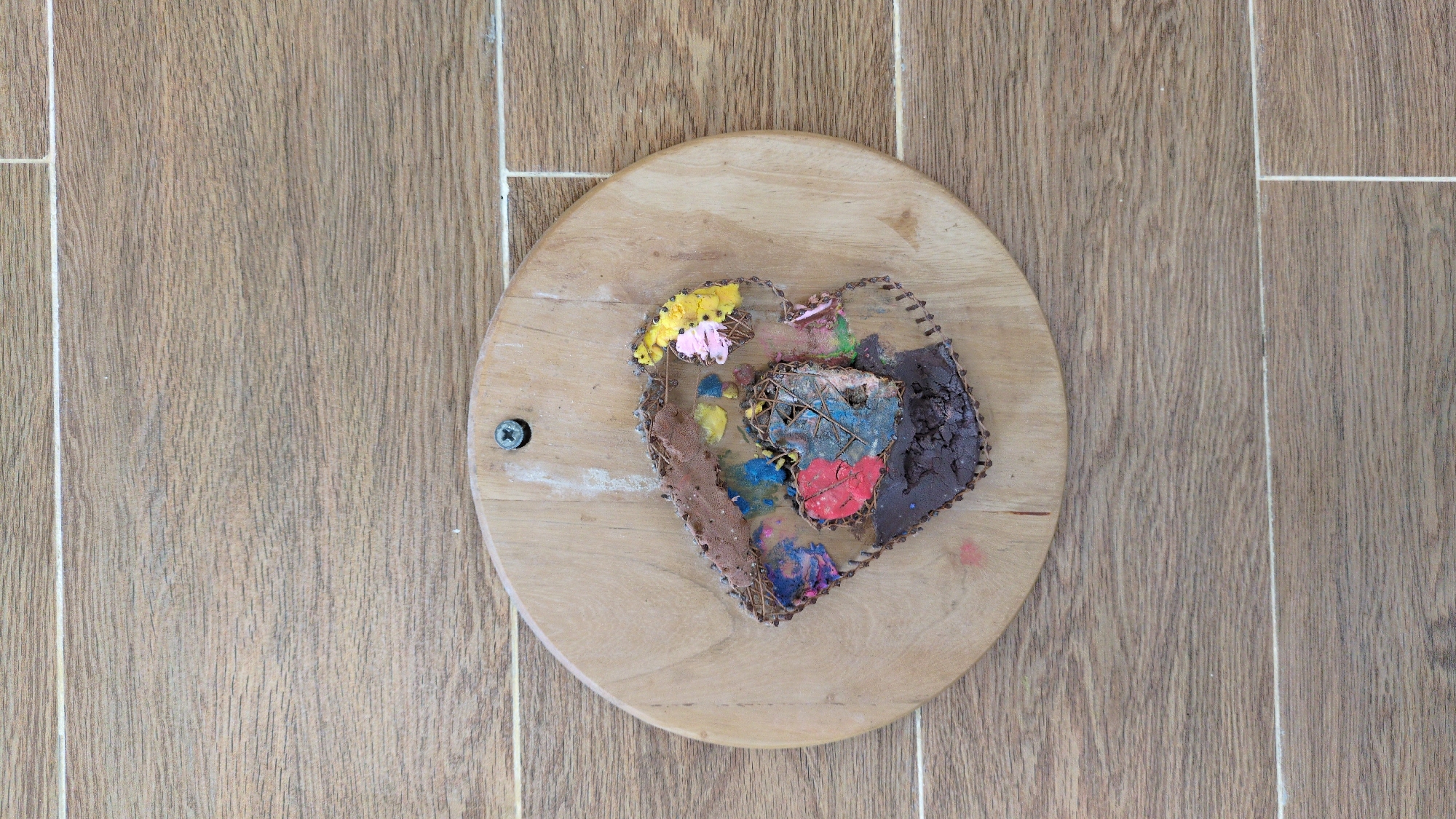 A top-down, eye-level shot shows a heart-shaped, mixed-media sculpture mounted on a light-colored wooden disk, which is secured to a wooden-tiled floor. The floor is composed of narrow, light brown wooden tiles with white grout lines. The sculpture consists of various materials including dried flowers (yellow and pink), brown textured material, and pieces colored with blue, yellow, red, and brown paint. Some of the pieces appear to be made of cardboard. The heart's perimeter is defined by small, dark-colored tacks. A single screw is visible on the lower left edge of the wooden disk. The location is plausibly Da Lat, Vietnam, based on the prevalence of similar wooden decor items.