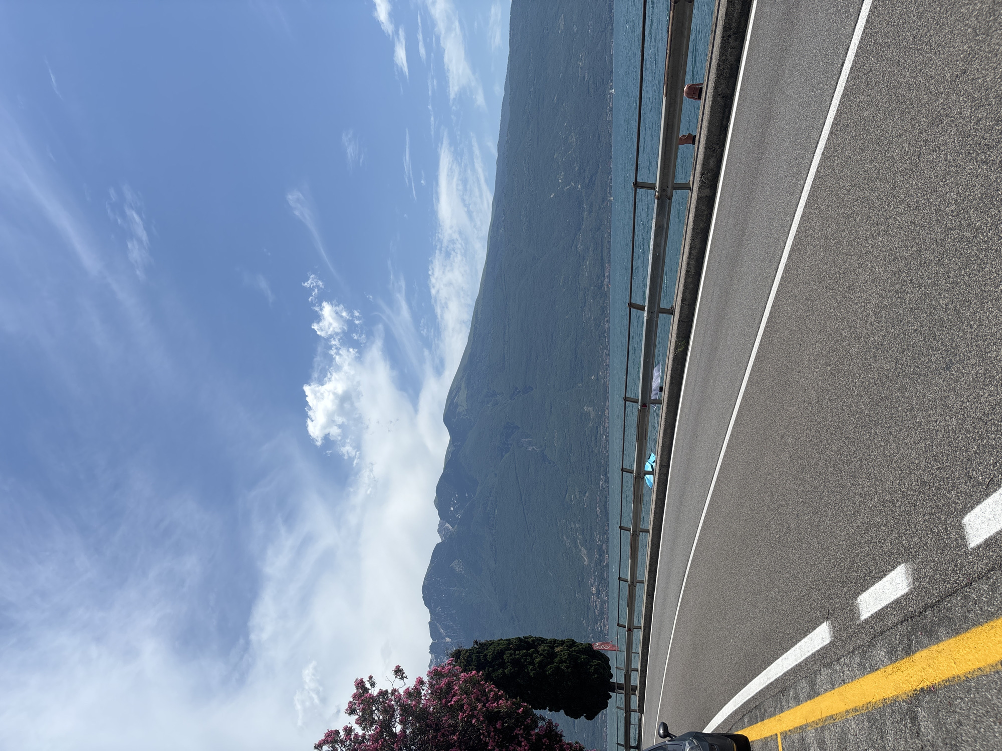 A paved road runs parallel to a large body of water, likely a lake, with a mountain range in the background. The road has white lane markings and a yellow line on the left edge. A metal guardrail separates the road from the water. On the water's edge, there are some people, and a blue object is visible. In the foreground left, a dark colored car is partially visible, and a tree with pink flowers and a dark green cypress tree stand beside the road. The sky is blue with scattered white clouds. The scene is likely located in Tignale, Italy, given the mountainous terrain adjacent to a lake.