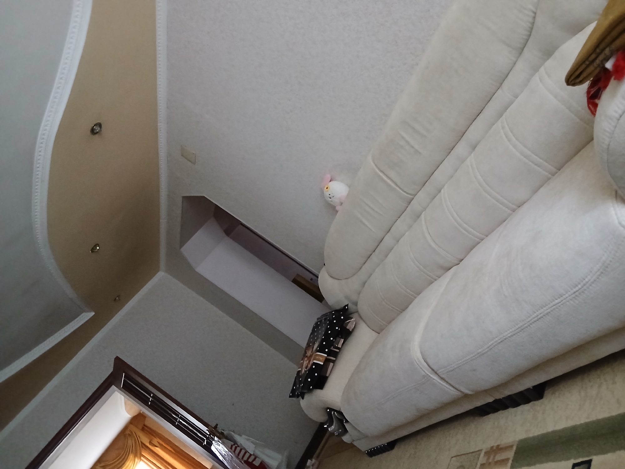 The image depicts an interior scene, likely a living room, with a large, cream-colored sectional sofa occupying a significant portion of the frame. A black and white polka-dotted box with a bow is placed on the armrest of the sofa. To the right of the sofa, a small, white stuffed animal with pink ears and eyes is visible. The walls are painted a light, textured color, and the ceiling features recessed lighting and decorative molding. A window with draped golden curtains is visible in the upper left corner. The image's perspective is angled, giving a sense of depth to the room. The presence of decorative elements suggests a domestic or hospitality setting. The image is of an interior space located in the city administration of Taraz, Kazakhstan.