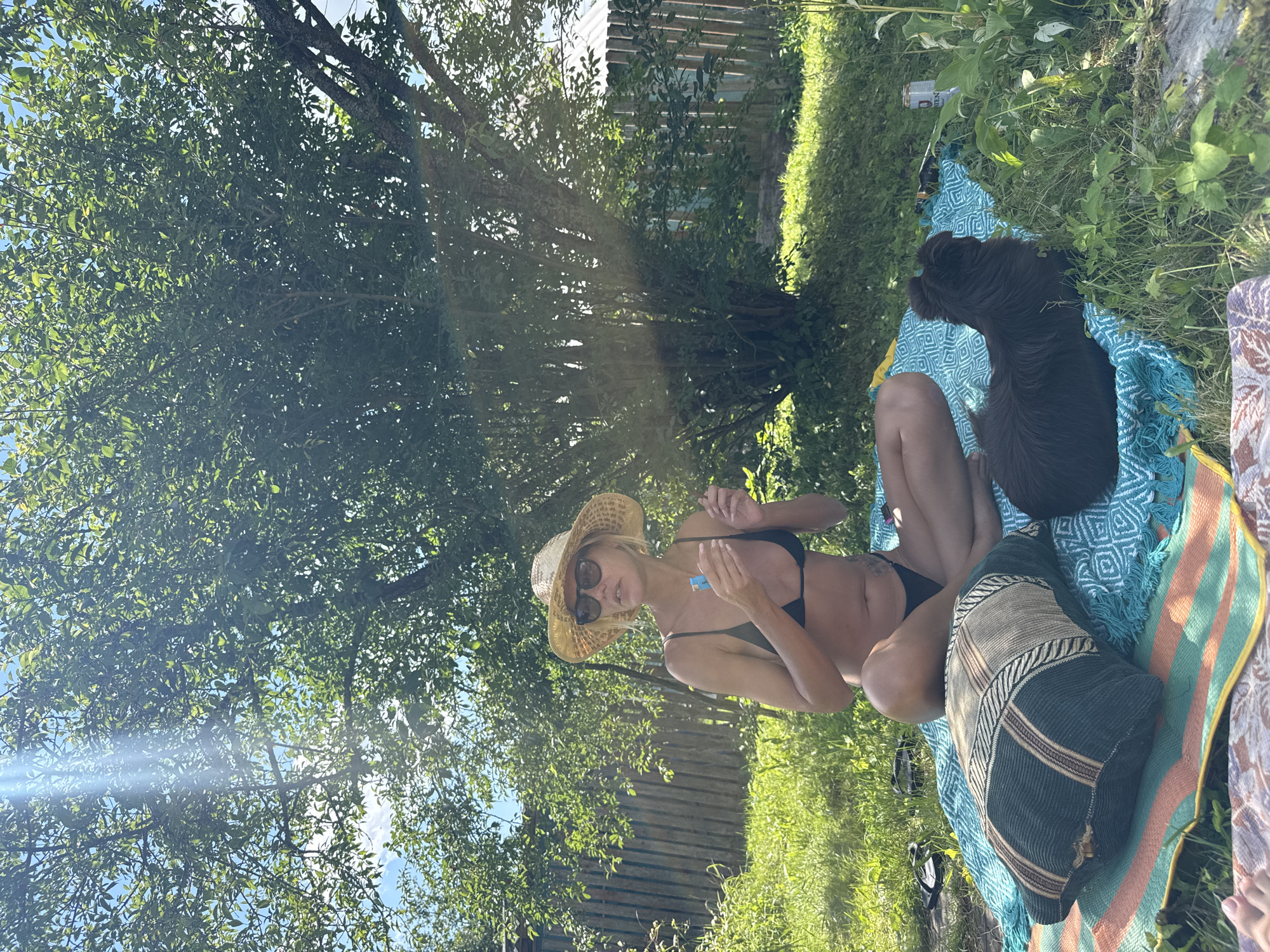 A woman wearing a black bikini and a straw hat is sitting on a blanket in a grassy yard, holding a blue object. Next to her is a dark brown dog. A wooden fence is visible in the background, and a large tree with green leaves casts a shadow over the scene. Sunlight streams through the leaves, creating lens flare. The location is identified as Krasnaya Presnya SNT, Russia.