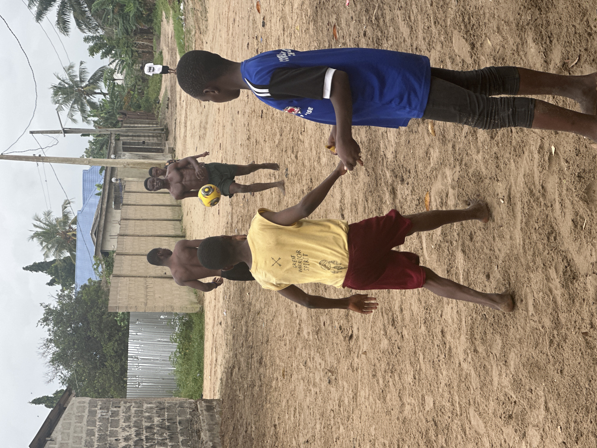 In Badagry, Nigeria, on a cloudy day, four young boys are playing soccer on a dirt field. Two boys are in the foreground, one facing away from the camera in a yellow t-shirt and red shorts, the other facing left in a blue t-shirt and dark leggings. In the middle ground, two other boys are more engaged with the soccer ball; one is kicking it, wearing dark shorts, and the other is walking towards the left. The environment is rural, with low-rise buildings and palm trees visible in the background. A corrugated metal fence and a concrete block wall are also present. The ground is sandy and uneven.