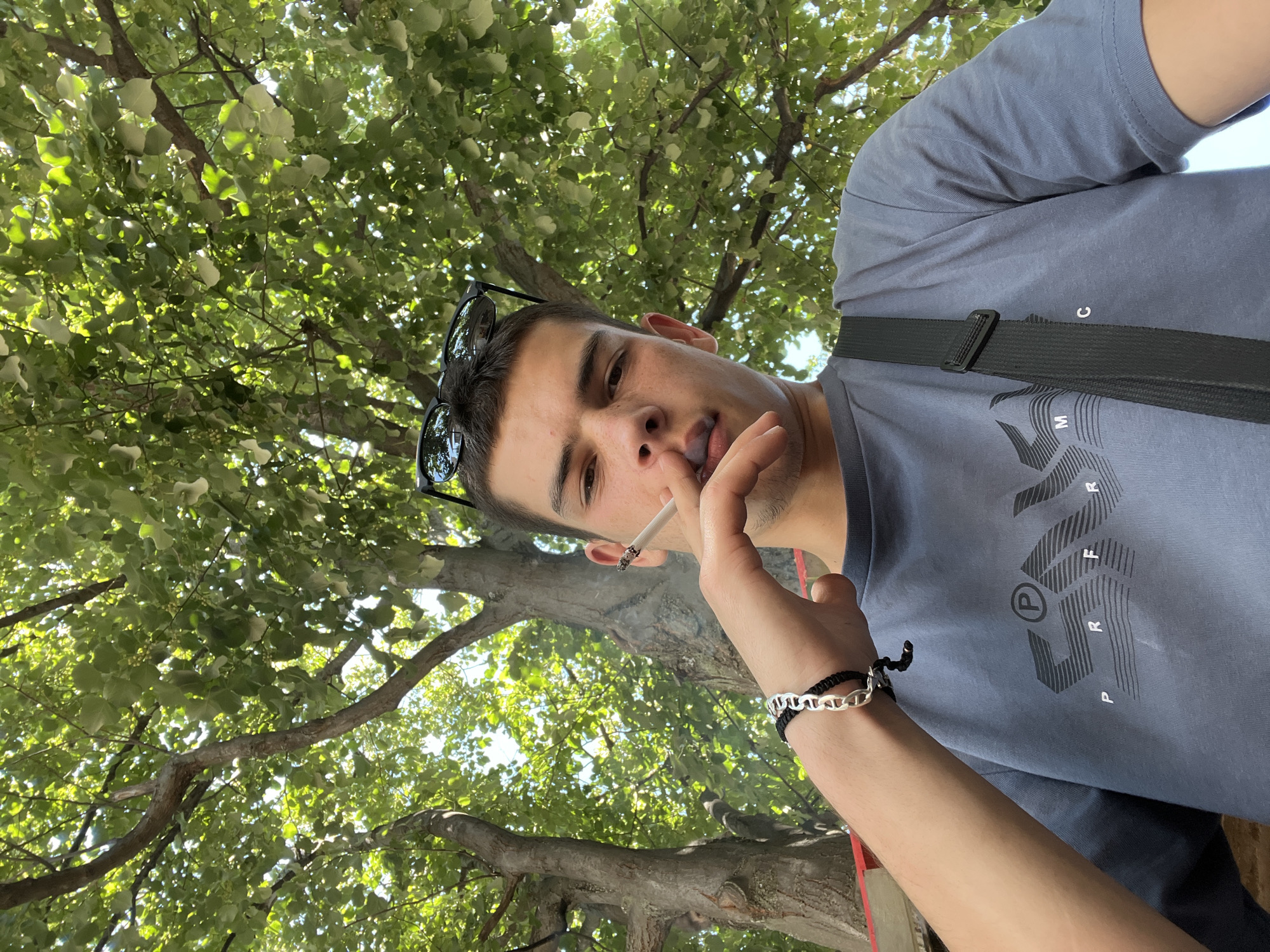 A young man is captured in a selfie from a low angle, with the canopy of a large tree and sunlight filtering through its leaves forming the background. He is wearing a grey t-shirt and has a black strap from a bag across his chest. He holds a lit cigarette between his fingers, bringing it to his lips, and appears to be exhaling smoke. A black bracelet with silver chain links adorns his left wrist. Sunglasses are perched on top of his head. The image appears to have been taken outdoors, possibly in a park or garden setting, given the prominent trees. The location is not specified as Sopot, Bulgaria.