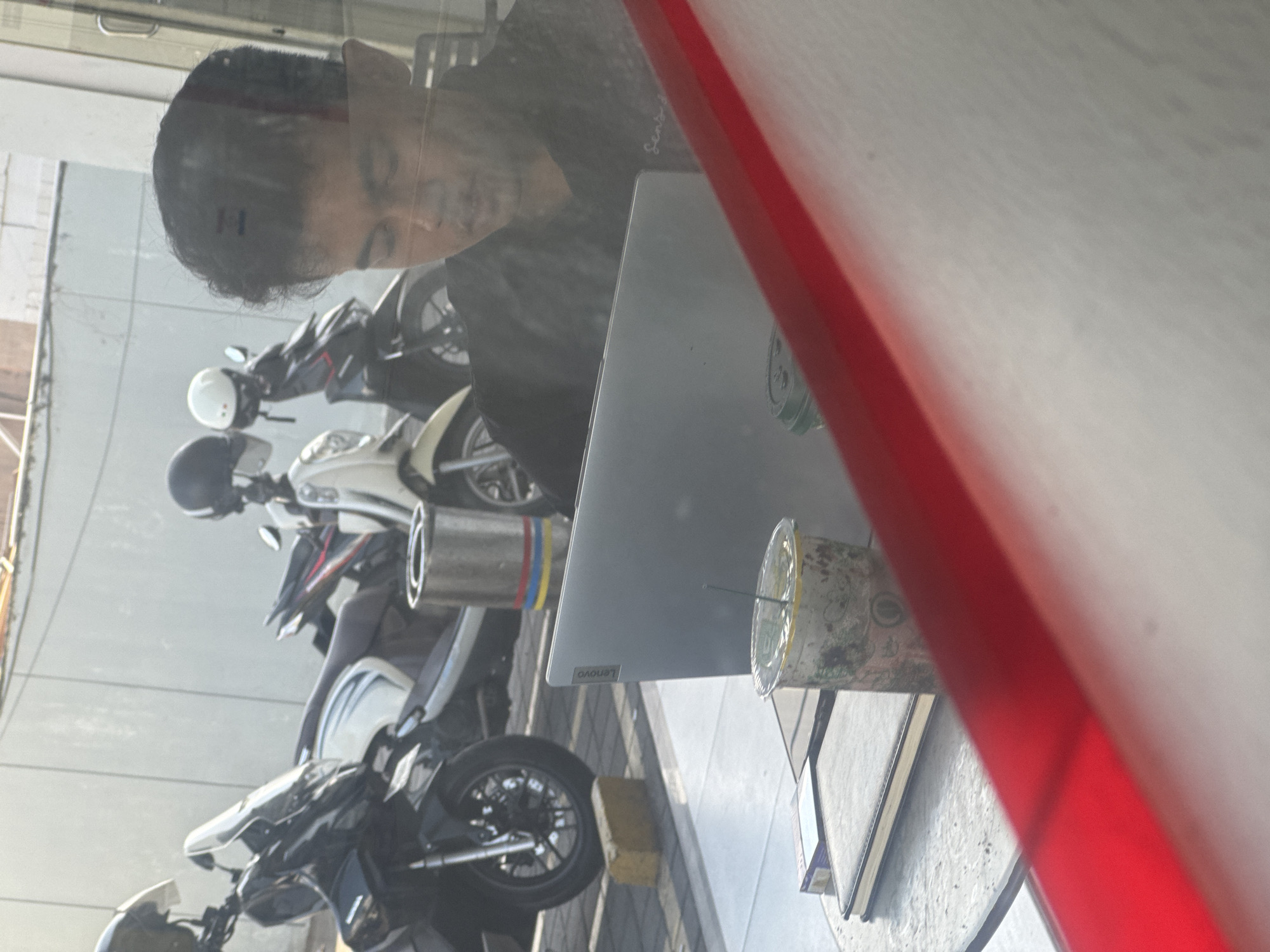 A medium shot captures a person sitting at a table with a laptop, reflected in a window. The person, with dark, curly hair and wearing a black shirt, is looking down at the laptop which is open on the table. To the left of the laptop is a clear plastic cup with a straw and a beverage inside, appearing to contain ice and some sort of green infusion. Beneath the cup and laptop are what seem to be notebooks or folders. In the background, several motorcycles are parked. The scene suggests an outdoor or semi-outdoor cafe or seating area, potentially near a street given the presence of parked motorcycles and the general urban aesthetic. The light indicates it is daytime, with sunlight casting shadows and highlighting the reflective surfaces. The overall impression is one of a casual, everyday moment, possibly in a bustling urban environment.