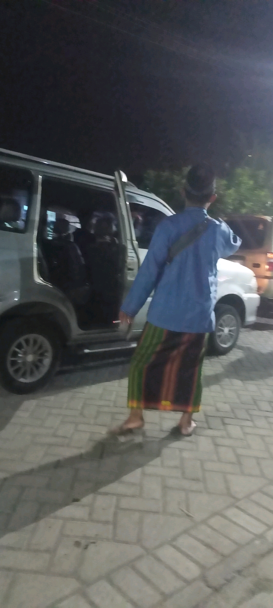 In Gempolklutuk, Indonesia, a man wearing a blue shirt and a sarong with green, brown, and orange stripes stands on a paved area next to an open-doored silver van. He is facing away from the camera, with his right arm bent and extended slightly. A dark-colored bag is slung across his left shoulder. The van's interior is visible, with multiple figures seated inside. Behind the van, a white vehicle is partially visible. The ground is made of interlocking light-colored pavers, and shadows are cast by the vehicles and the man. The scene appears to be outdoors at night, with some ambient lighting.