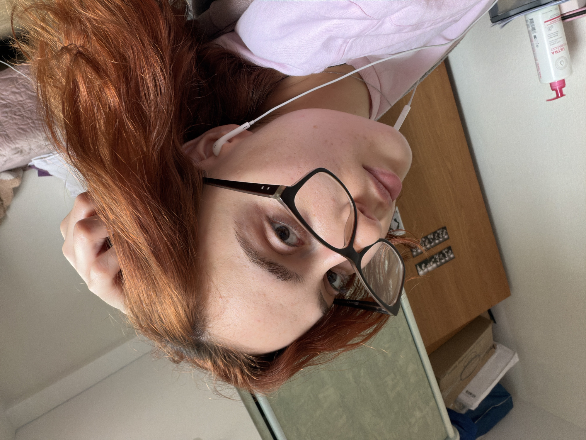 The image, originally rotated 90 degrees clockwise, captures a close-up, low-angle portrait of a person indoors. The subject, likely a young adult, has long, reddish-brown hair with visible darker roots and wears black-framed, rectangular glasses. They are looking directly at the camera with a neutral expression.
The person is wearing white wired in-ear headphones in their right ear and a light pink top, possibly a hoodie. Their skin appears clear with visible freckles. The background features a light wooden surface, likely a desk or table, positioned to the person's right side, and a plain white wall behind it.
On the wooden surface, a white bottle with a red flip-top cap is visible. The label clearly shows the French text "ULTRA HYDRATANT," with "Topicrem" also partially visible, consistent with the given location context of Strasbourg, France. Small, dark rectangular objects, possibly decorative or organizational items, are arranged in rows on the wooden surface. Below this surface, various boxes and other items are stacked. The bright, even lighting suggests it is daytime. The scene conveys a casual, relaxed indoor moment.