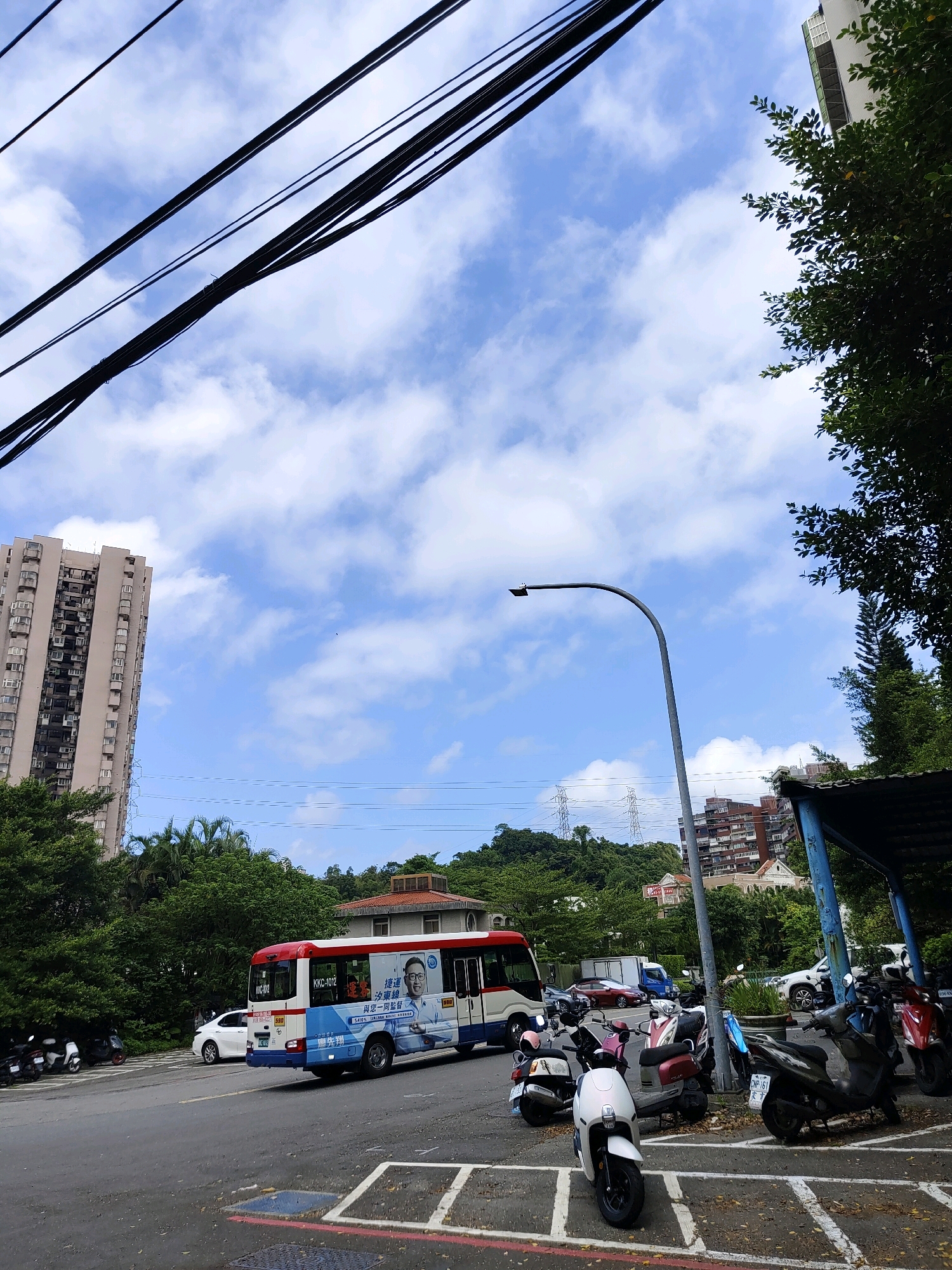 The image captures a street scene in New Taipei City, Taiwan, under a bright sky with scattered clouds. Dominating the upper left are thick, black electrical cables strung across the sky, typical of urban environments. To the left, a tall, beige apartment building with many windows stands against the blue sky. In the foreground, a paved road shows white parking lines and a few fallen leaves. A red and white bus, with Chinese text and an advertisement featuring a man's portrait, is driving on the road. Adjacent to the road, a row of parked motorcycles is visible. Behind the bus, a white car is also parked. The background features lush green trees, more residential buildings of varying heights and architectural styles, and rolling hills with power transmission towers. A modern street lamp with a curved pole is positioned in the mid-right of the frame. A blue awning structure is also present on the right side of the image.