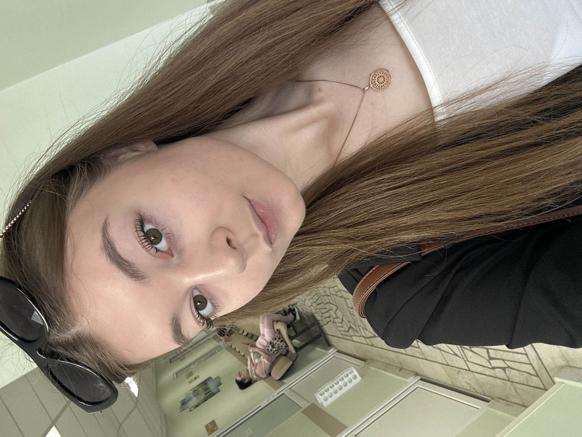 The image captures a selfie of a young woman in an indoor setting in Kazan, Russia, rotated 90 degrees counter-clockwise.
The primary subject is a young woman with long, straight, brown hair that frames her face and extends beyond the frame. She has light-toned skin, dark brown eyes accented with mascara, and light pink lips. She wears a white top, partially visible, layered under a black jacket or garment. A delicate gold-toned necklace with a circular, intricately patterned pendant, possibly a sun wheel or mandala, is prominently displayed on her neck. Black sunglasses are perched on the left side of her head, resting above her ear. A brown strap, likely from a shoulder bag, is visible over her right shoulder. Her gaze is direct and her expression is calm and neutral.
In the background, the setting appears to be a public interior space, such as a waiting room or hallway in a clinic or office building. The floor is covered with light-colored tiles featuring a distinctive repeating geometric pattern. Light-colored walls and what appear to be doors or partitions with glass panels are also visible. Further back, another person is seated on a bench or chair, partially obscured, suggesting they are also waiting. There are some indistinct rectangular shapes on the wall that could be posters or notices. The lighting is bright and even, typical of well-lit indoor environments during the day.
The main activity is the woman taking a selfie. No clearly legible text is visible on any signs, labels, or objects in the background.