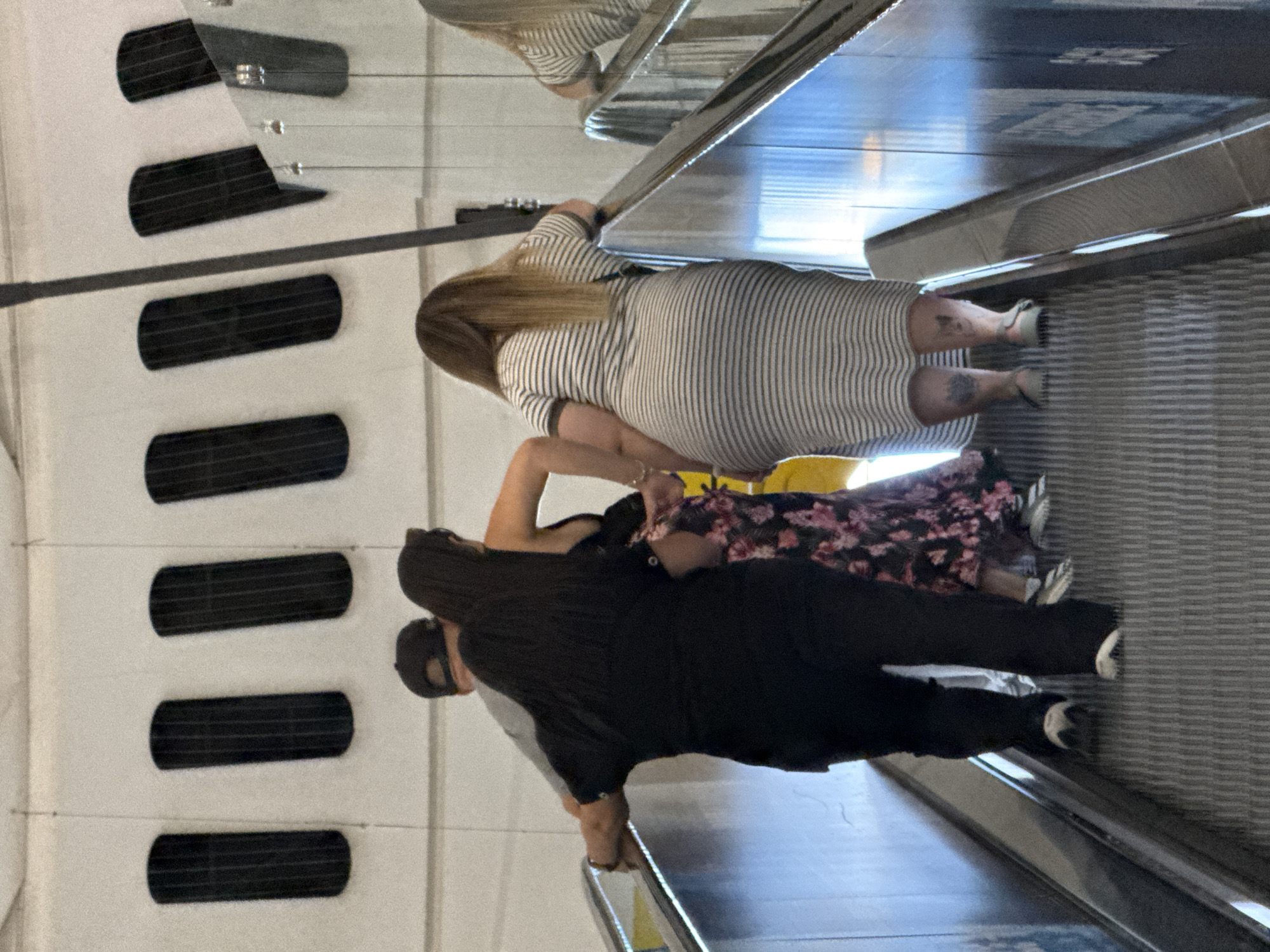 The image shows a group of four people on an escalator. The escalator is moving upwards. The people are dressed in casual attire. Two women are in the foreground, one wearing a black outfit and the other a striped dress. Behind them, a man in a grey t-shirt and a black cap is visible, along with another person partially obscured. The background shows a white wall with vertical black decorative elements. The escalator handrails are metallic, and the steps are ribbed. Reflections of the surrounding environment are visible on the metallic surfaces of the escalator. The scene appears to be in an indoor public space, possibly an airport or a shopping mall, and there is no clear indication of the location being in Barcelona, Spain.