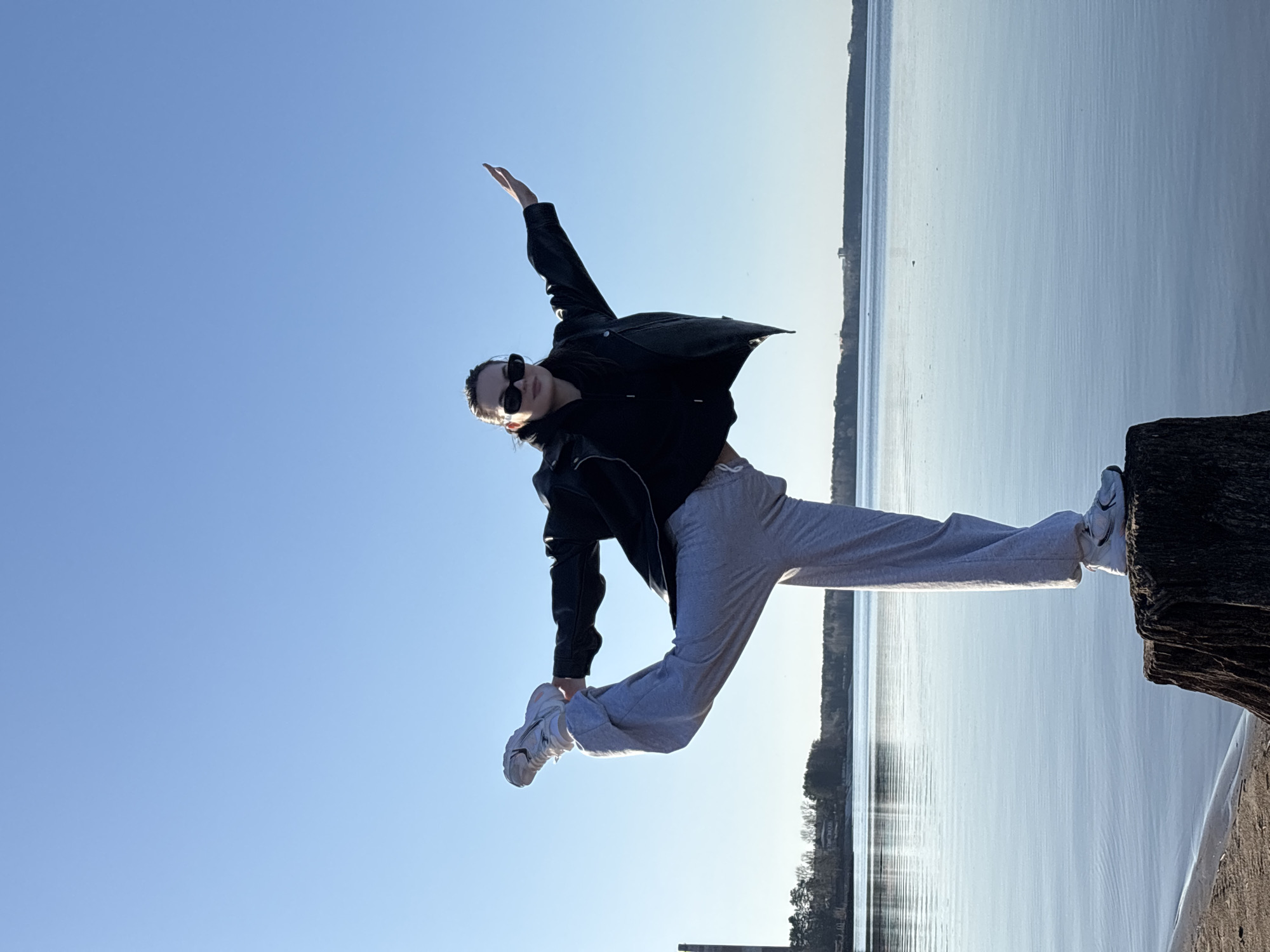 A person in athletic attire, wearing sunglasses and a black jacket over gray sweatpants and white sneakers, performs a split in front of a body of water. The person is balanced on a wooden structure, possibly a pier or breakwater. The background shows a shoreline with trees and buildings. The sky is a clear, light blue. The location is likely in Chaikovsky, Russia, based on the presence of the Kama river in the background, which is a feature of this city. The sun is bright.