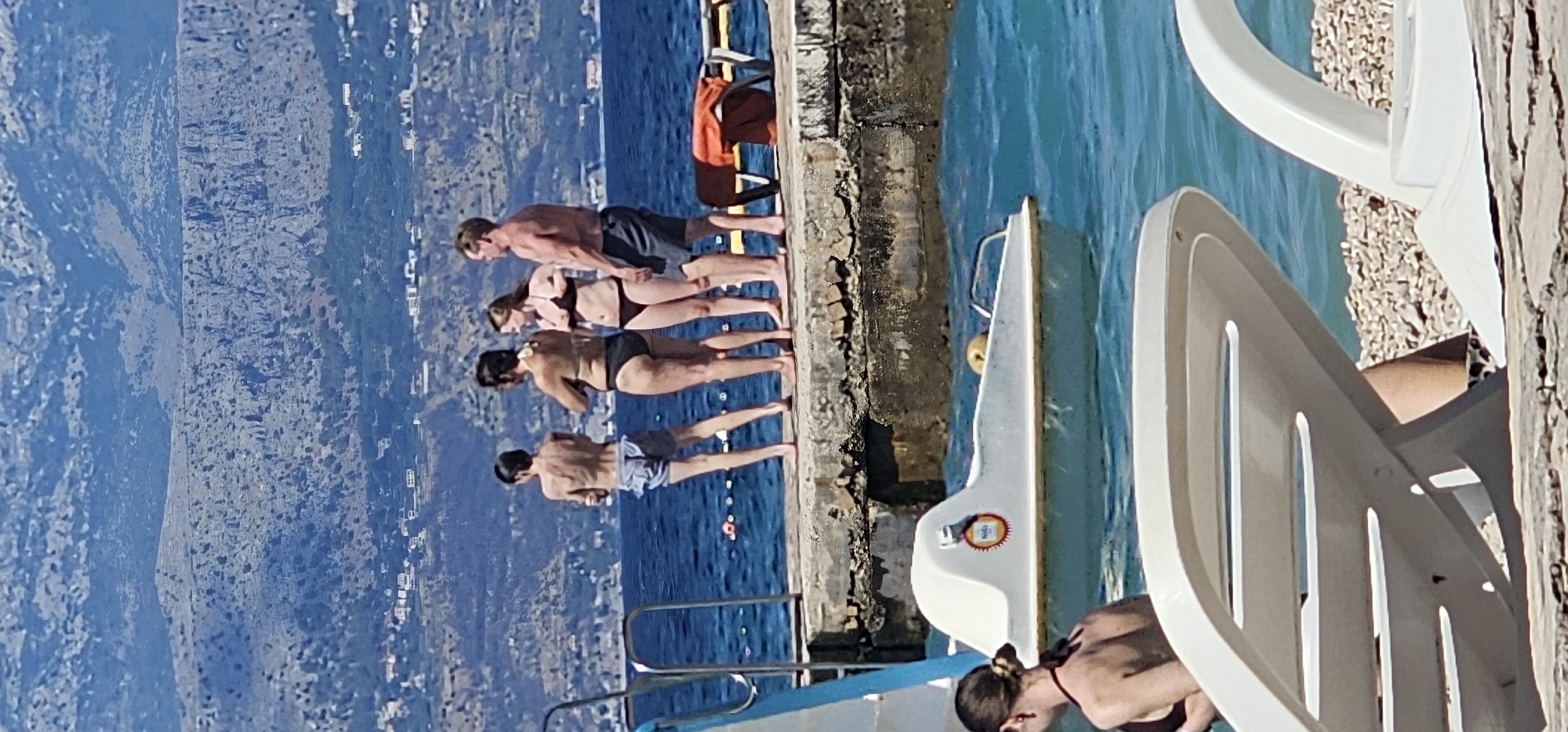 A group of four people stand on a concrete pier overlooking the Adriatic Sea in Supetar, Croatia. The two individuals on the left are wearing swim shorts and denim shorts, respectively. To their right, two women are dressed in bikinis, with one woman looking to her right and the other looking forward. A man in swim shorts stands at the far right of the group, facing towards the sea. In the foreground, a white paddleboat with a red and yellow circular logo rests in the turquoise water. In the background, a hazy mountainous landscape with scattered buildings rises behind the sea. Two white lounge chairs are partially visible in the bottom foreground, and a small orange life preserver is on a wooden chair to the right of the group.