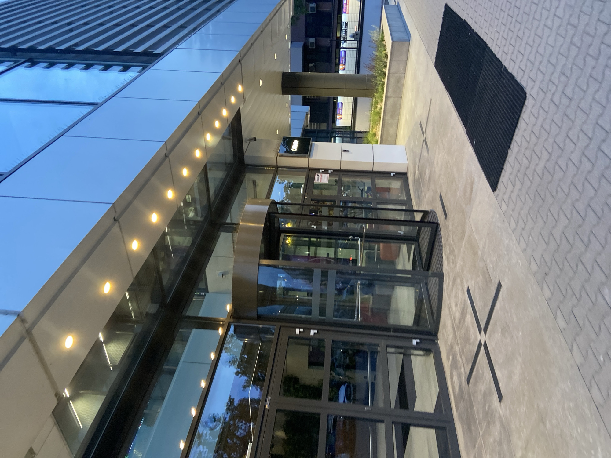 The image shows the modern glass and steel entrance of a building in Warsaw, Poland, likely an office building or hotel, given the contemporary architecture and signage. The entrance features a revolving door flanked by standard glass doors. Above the entrance, a contemporary overhang is illuminated by a series of recessed yellow lights. The building's facade is predominantly glass, reflecting the blue evening sky and surrounding structures. On the pavement in front of the entrance, there is a black cross-shaped marking and a long black mat. A planter box with low greenery is visible to the right of the entrance. In the background, there are signs on the building, including one that appears to read "CANAL+". The overall impression is one of a sleek, urban environment, likely captured during dusk or early evening.