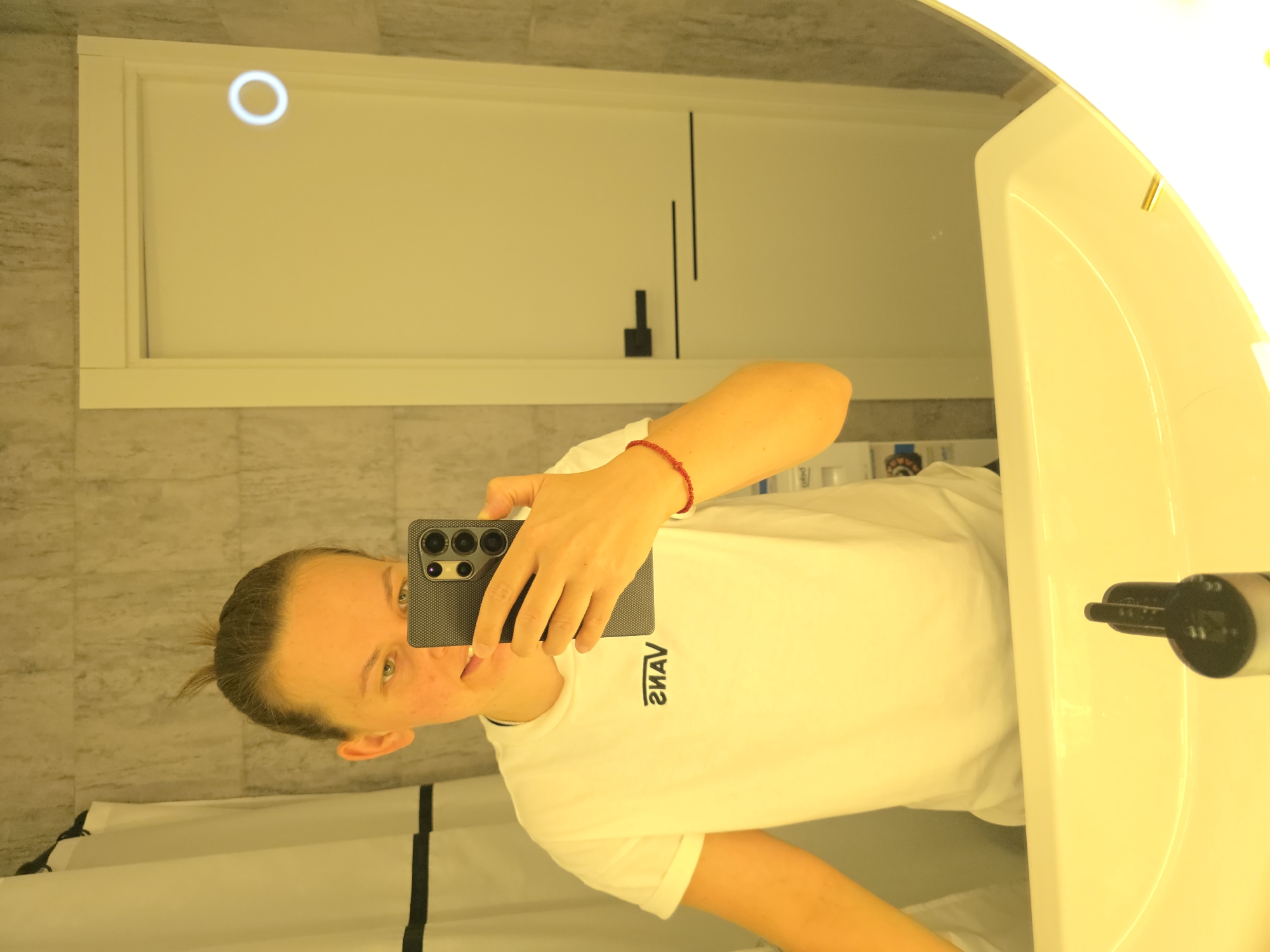 A young woman with her hair in a messy bun takes a selfie in a brightly lit bathroom. She is wearing a white t-shirt with a black Vans logo on it. She is holding a black smartphone in her right hand, and a red beaded bracelet is visible on her left wrist. The bathroom has gray tiled walls and a white door with a circular light fixture. A white shower curtain is visible on the left side of the image. A white sink is in the foreground, with a black soap dispenser on it. The lighting in the bathroom is very warm and yellow.
