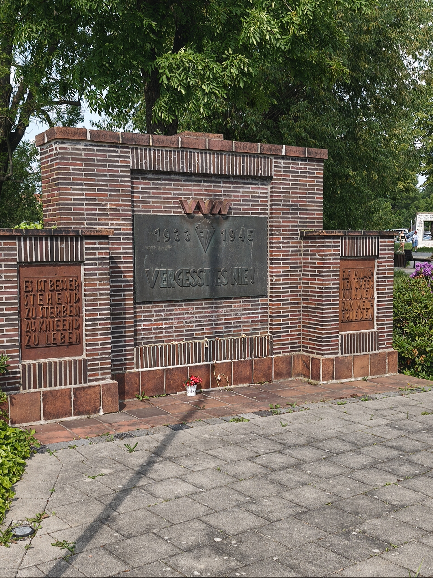 The image shows a war memorial in Unknown, Unknown. The memorial is constructed of red brick with darker brown accents. In the center is a bronze plaque with the dates "1933" and "1945" and the words "VERGESST ES NIE!" A red vase of flowers is placed in front of the memorial. To the left and right of the central plaque are smaller plaques with text in German. The memorial is surrounded by a paved area and greenery. In the background, there are trees and a glimpse of people and cars.