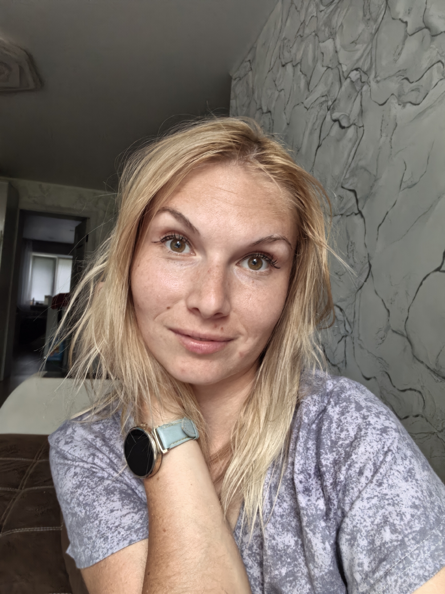 A young woman with blonde hair, freckles, and brown eyes looks at the camera. She is wearing a grey t-shirt with a speckled pattern and a light blue watch on her left wrist. She is indoors, in what appears to be a living room. The background includes a textured wall with a grey and white marble-like pattern, a doorway leading to another room, and a portion of a ceiling. The overall lighting suggests it is daytime. There is no indication this image is located in Sterlitamak, Russia.