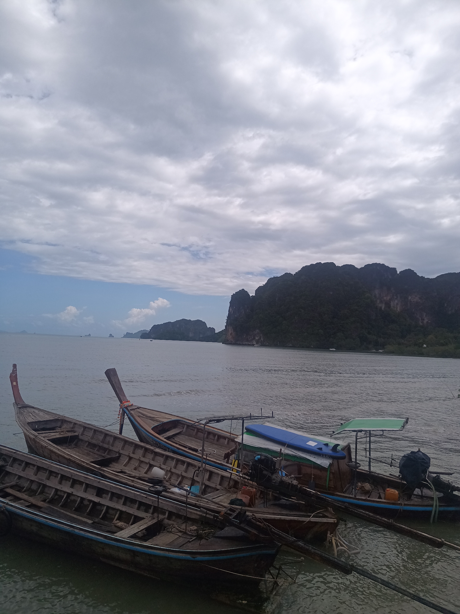 The image captures a coastal scene in Ao Nam Mao, Thailand. Three traditional wooden longtail boats are clustered near the shore, partially submerged in the water. The boats display details like the ribbed interiors and a blue surfboard resting on one. In the background, a body of water stretches out towards a mountainous coastline under a cloudy sky. Small islands or rock formations are visible in the distance.